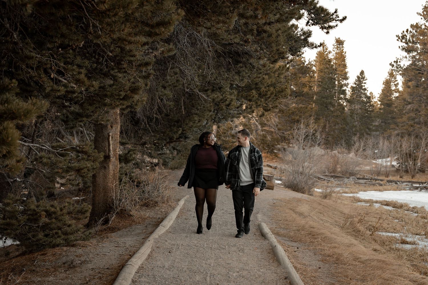 Couple walking hand in hand on a snowy forest path surrounded by pine trees in winter.