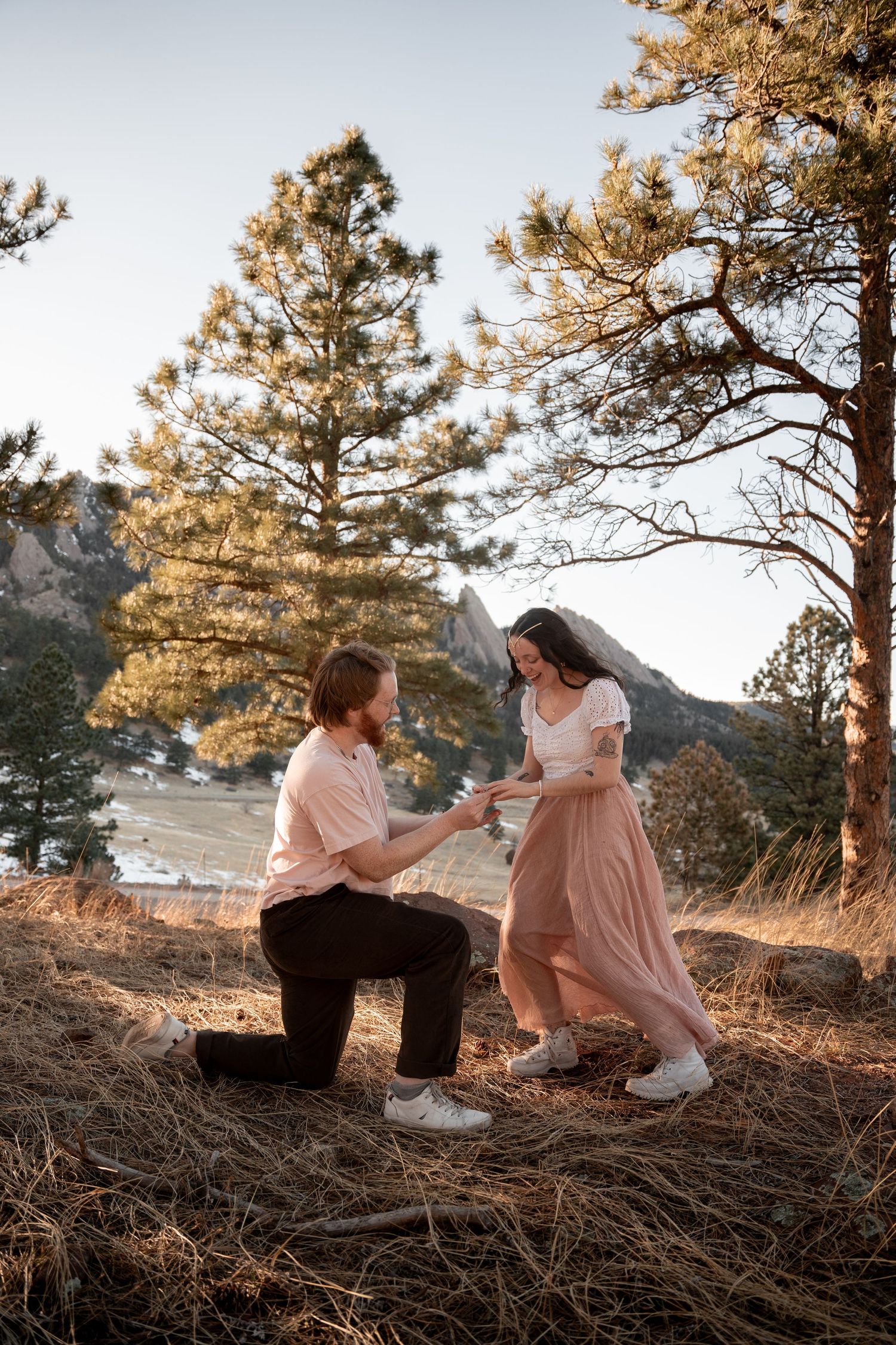 A romantic outdoor engagement photoshoot at sunset with a couple in pink attire among pine trees.