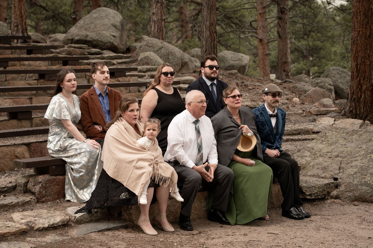 A family group poses together on stone steps surrounded by pine trees and boulders at an outdoor wedding celebration.