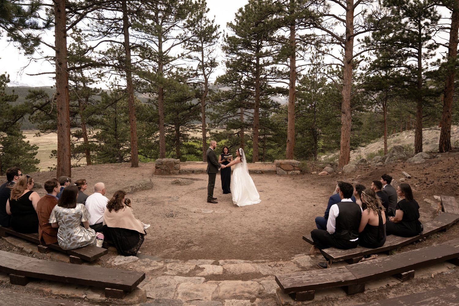 Outdoor wedding ceremony in a rustic pine forest amphitheater with guests seated on wooden benches surrounding the couple.
