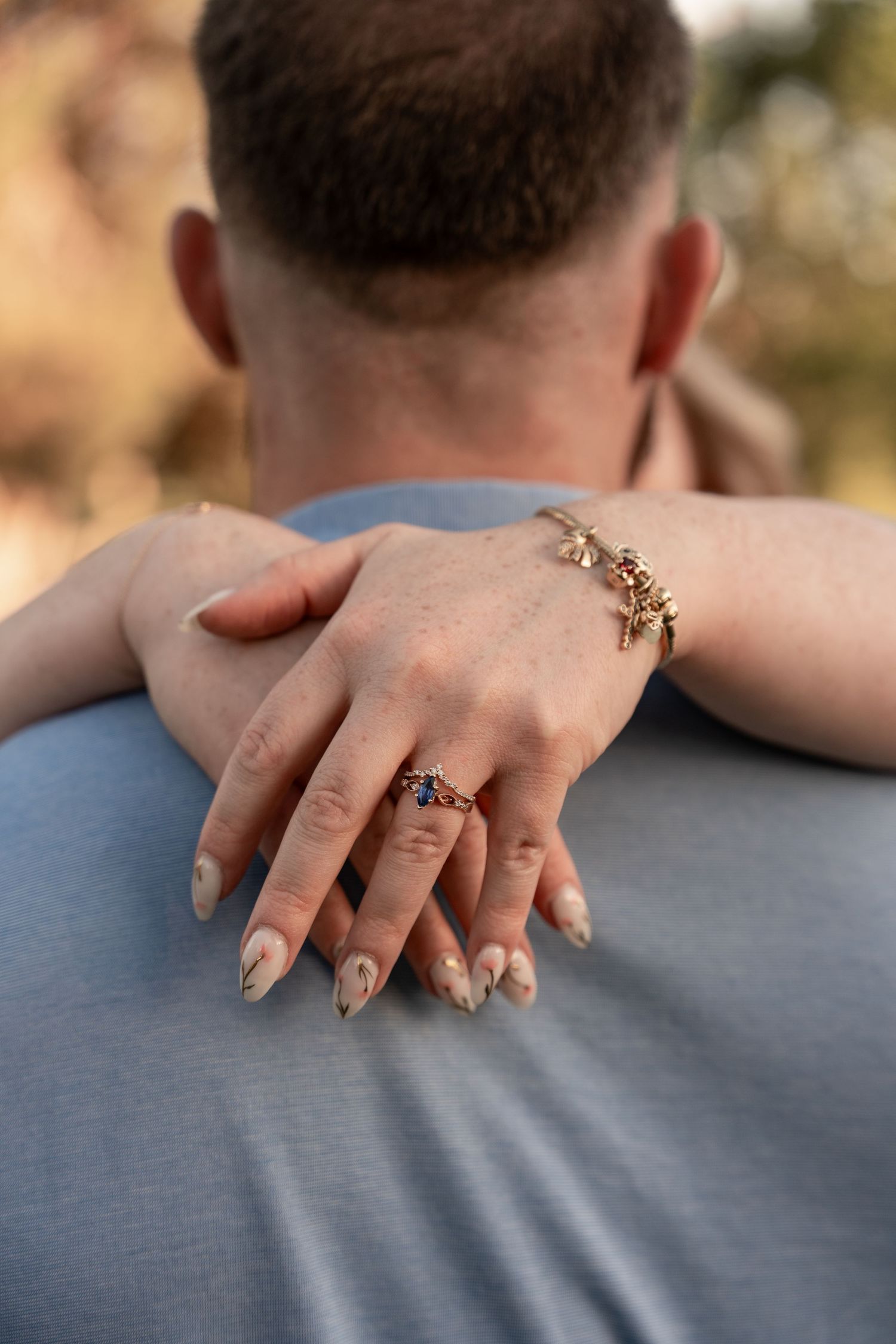 Woman's hand with gemstone ring and gold bracelet resting on man's shoulder, showcasing elegant nail art.