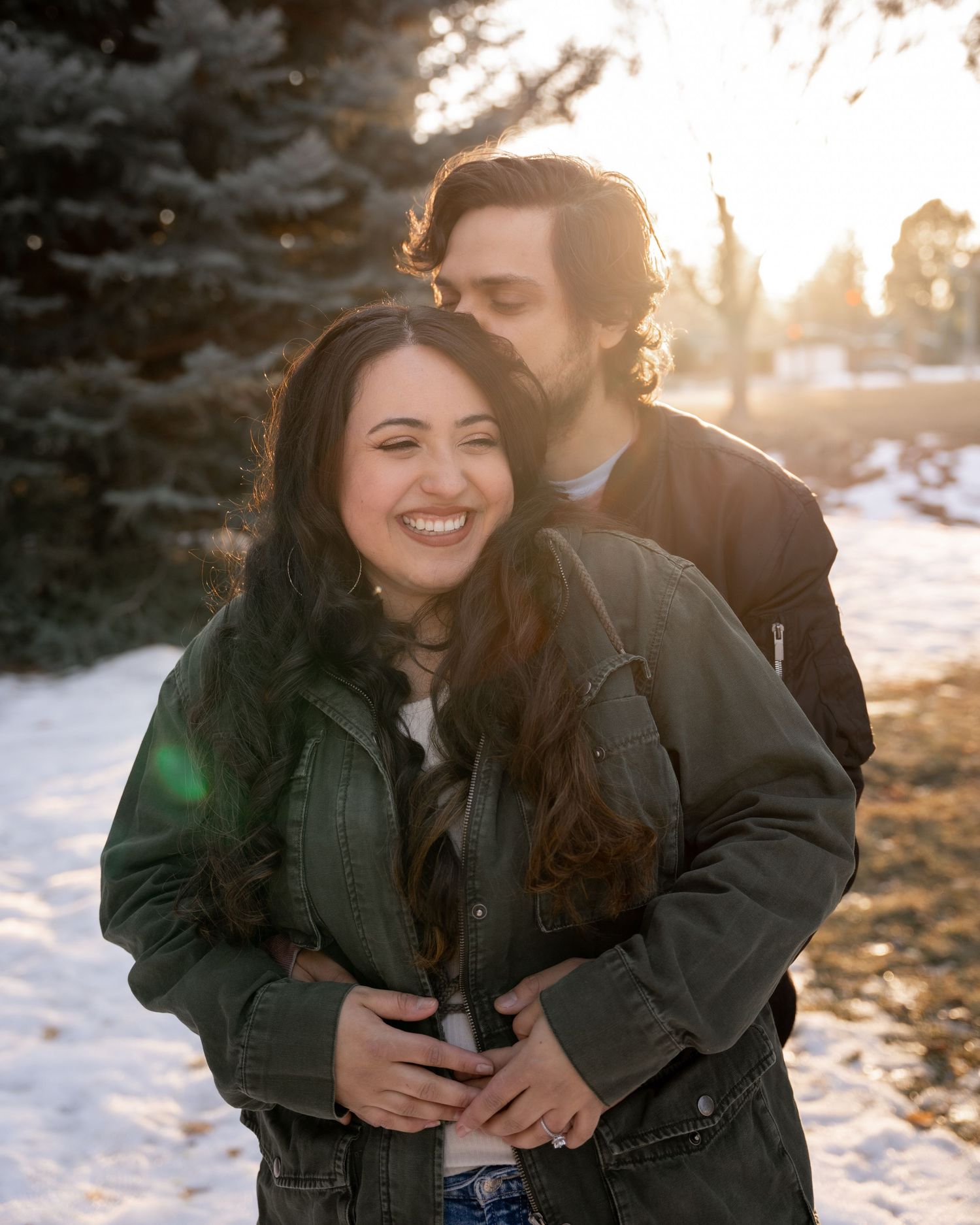 Couple embracing outdoors in snow, woman smiling as man kisses her head, golden sunset light, evergreen trees background.