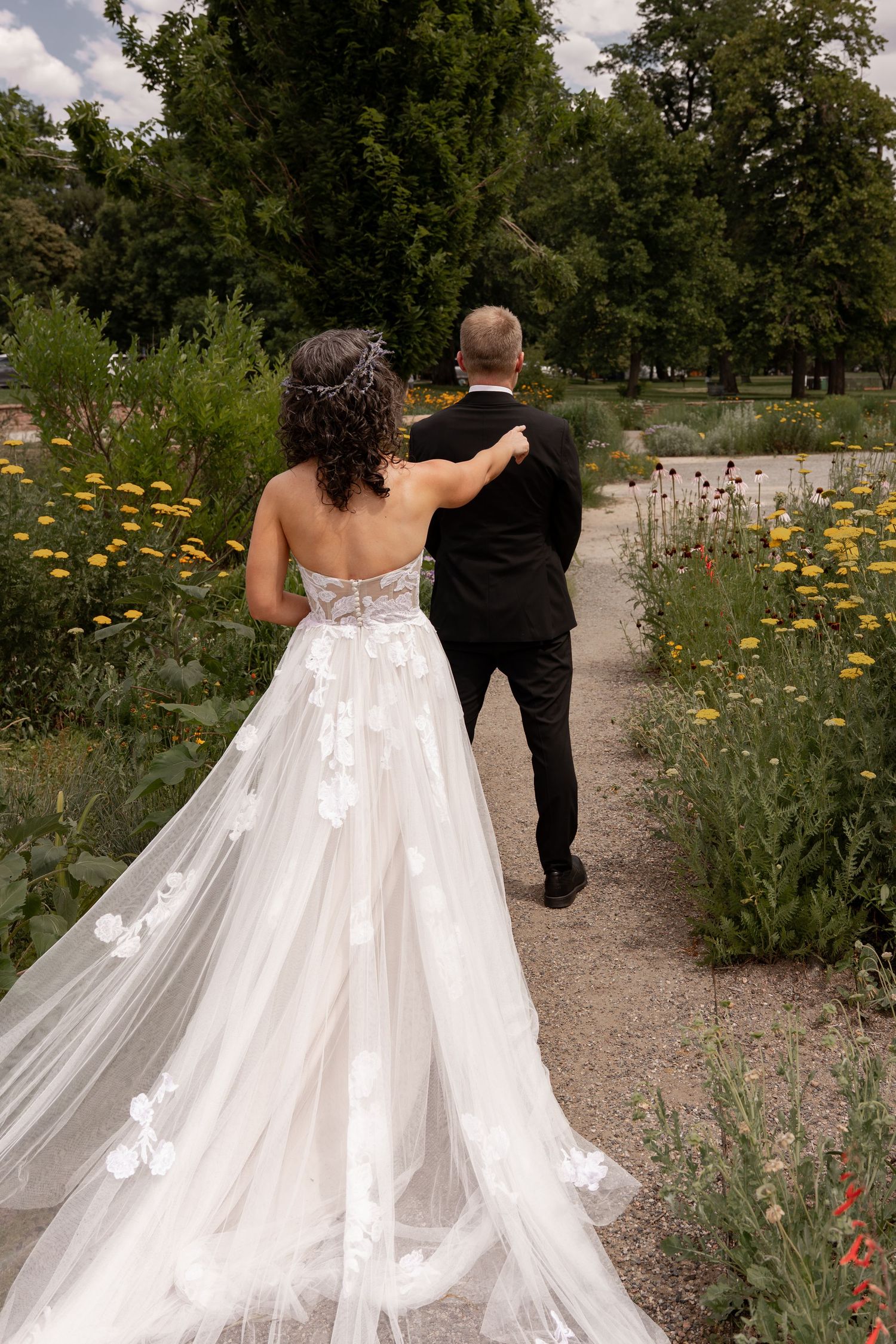 A couple in wedding attire walks through a garden path surrounded by wildflowers and trees on their special day.
