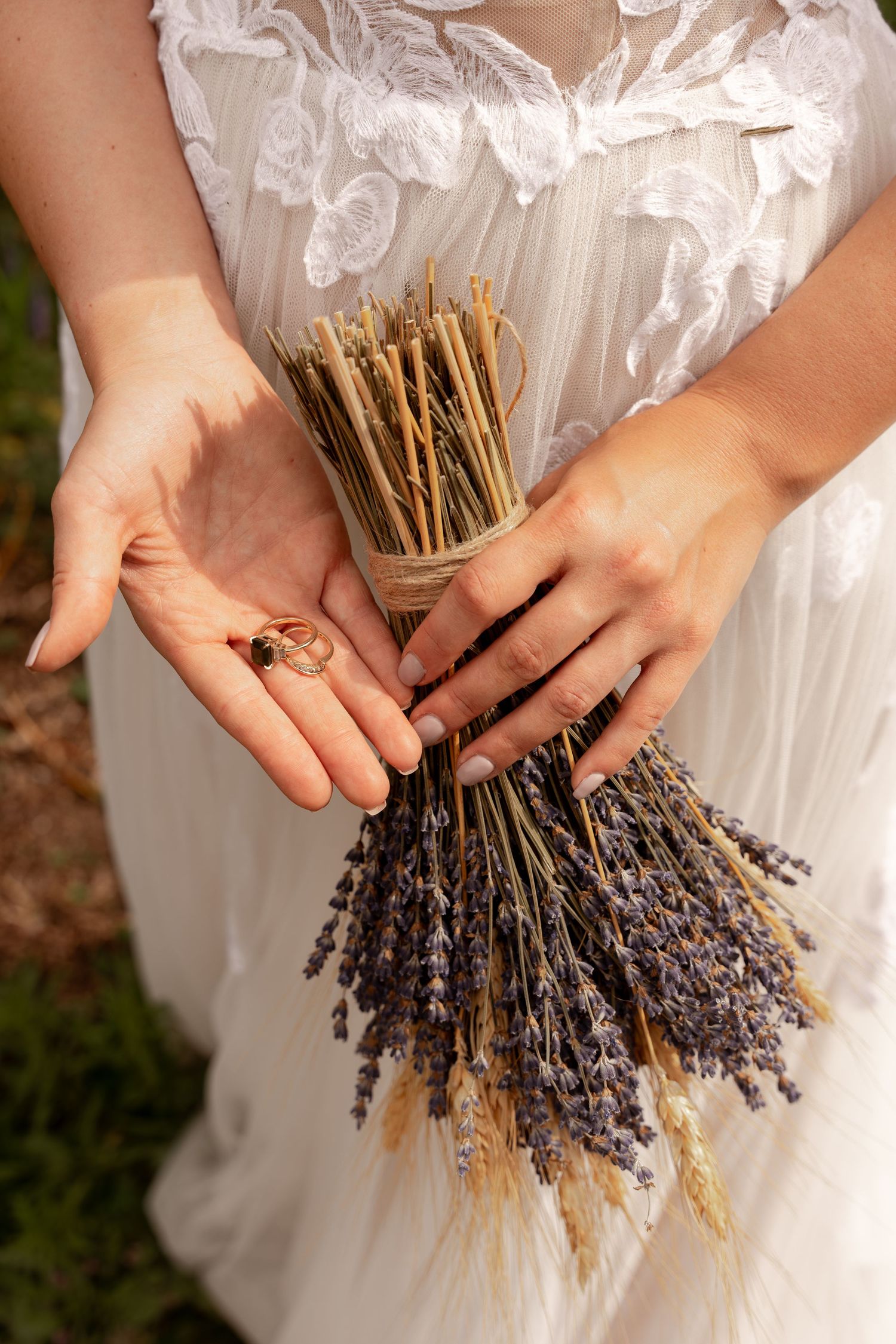 A rustic wedding bouquet featuring dried lavender and wheat stalks held in white-gloved hands.