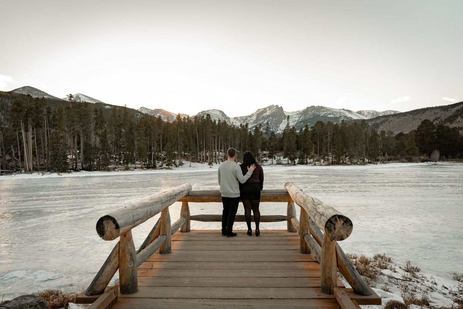 Couple embracing on wooden dock overlooking frozen lake with snow-capped mountains beyond.