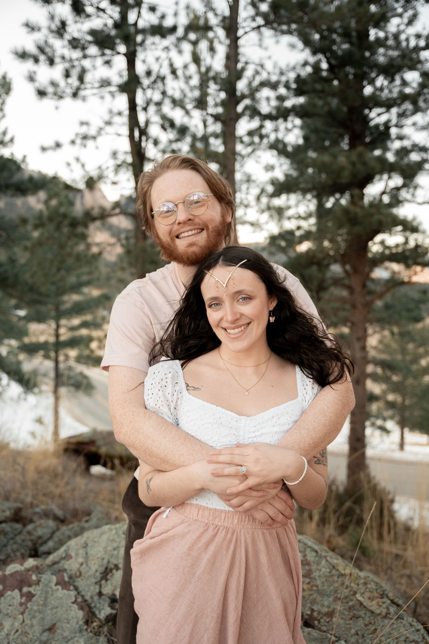 Smiling couple embracing outdoors among pine trees and rocky terrain, woman in white top and pink skirt.