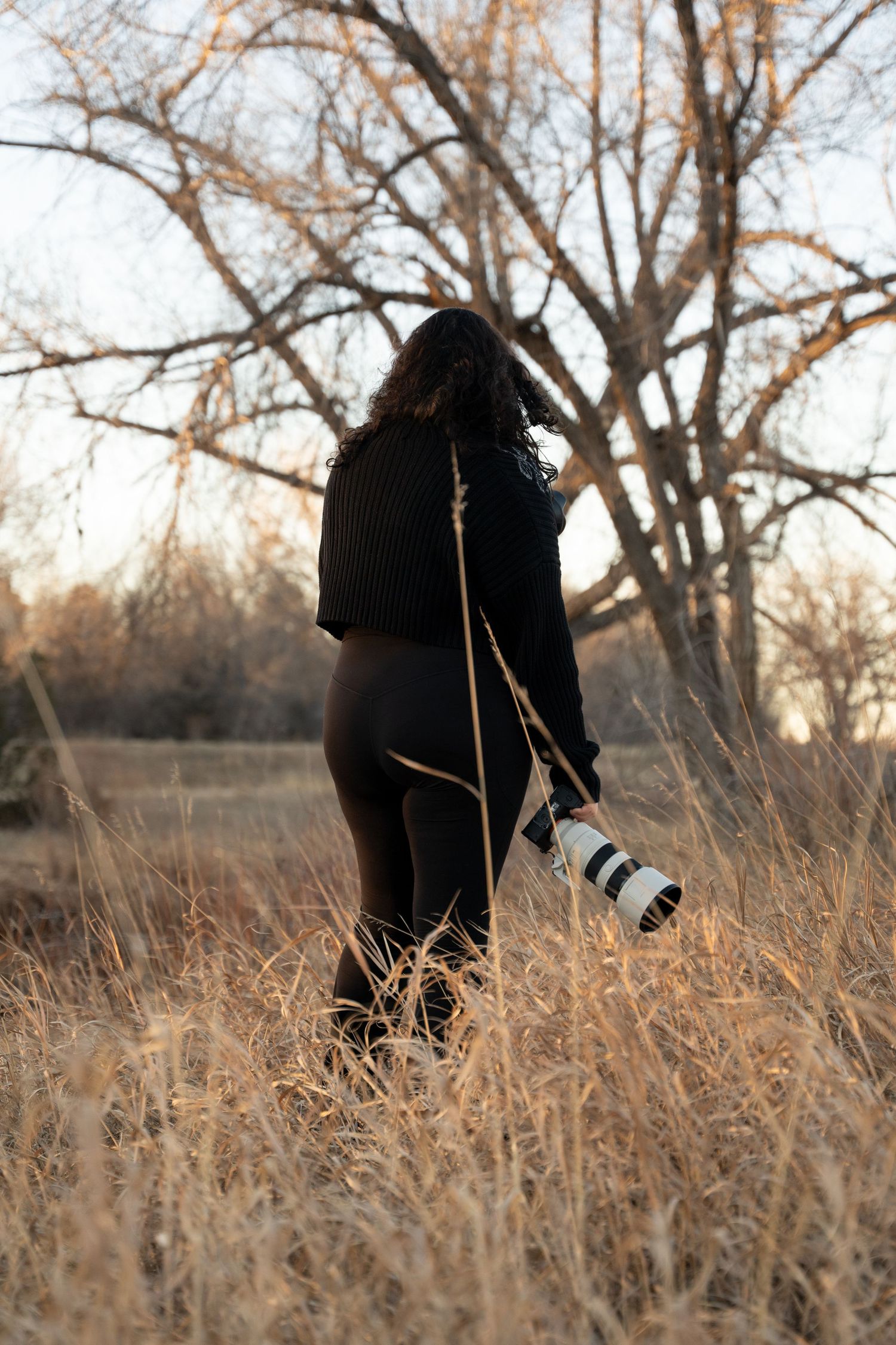 A photographer stands in dried grass holding a camera with telephoto lens at sunset.