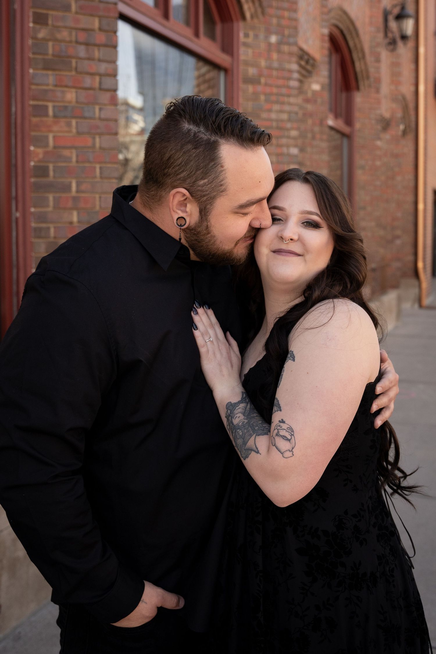 Couple in matching black outfits share a romantic moment against a brick building, woman smiling as man kisses her cheek.