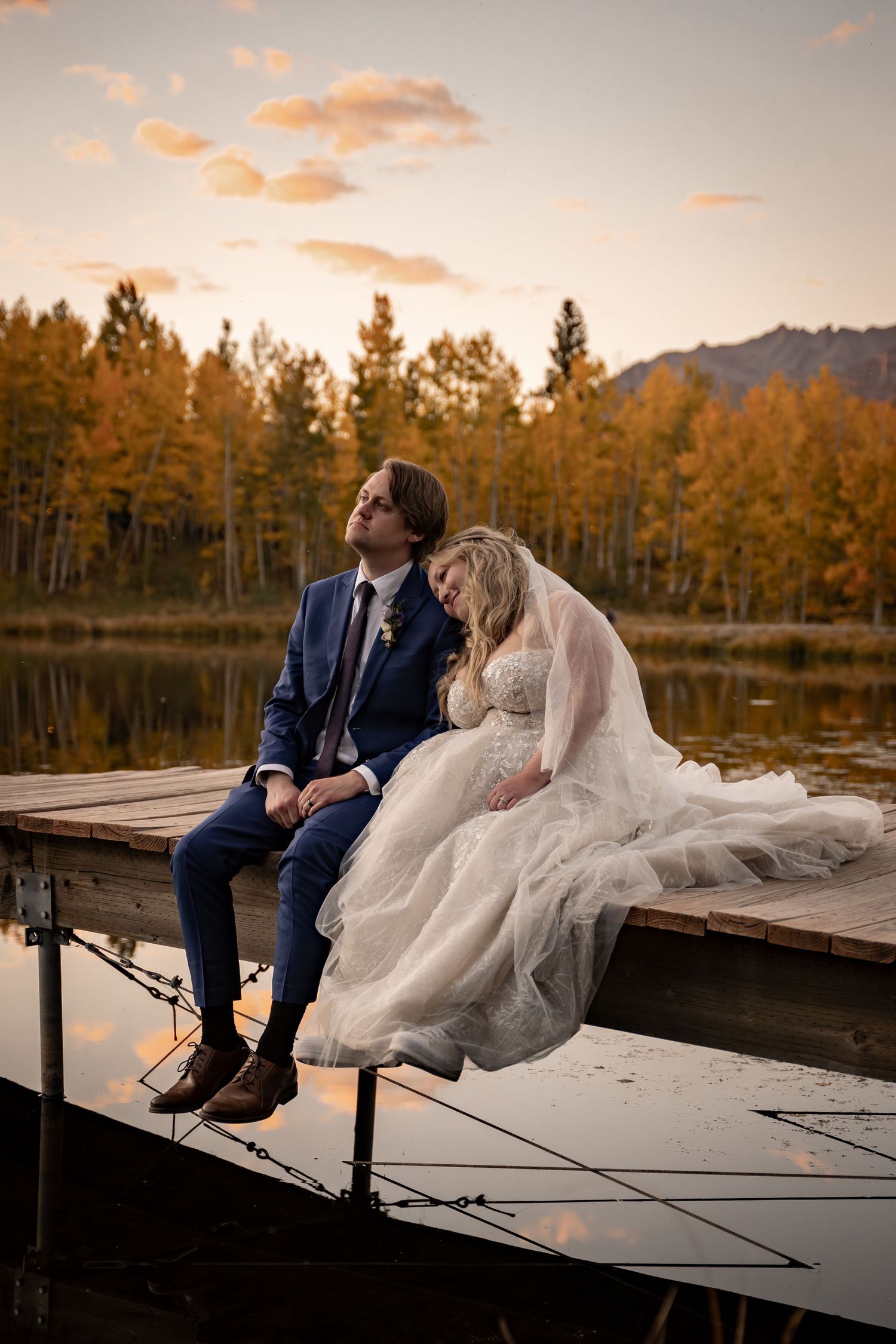 Newlyweds sitting together on a wooden dock at sunset beside a mountain lake.