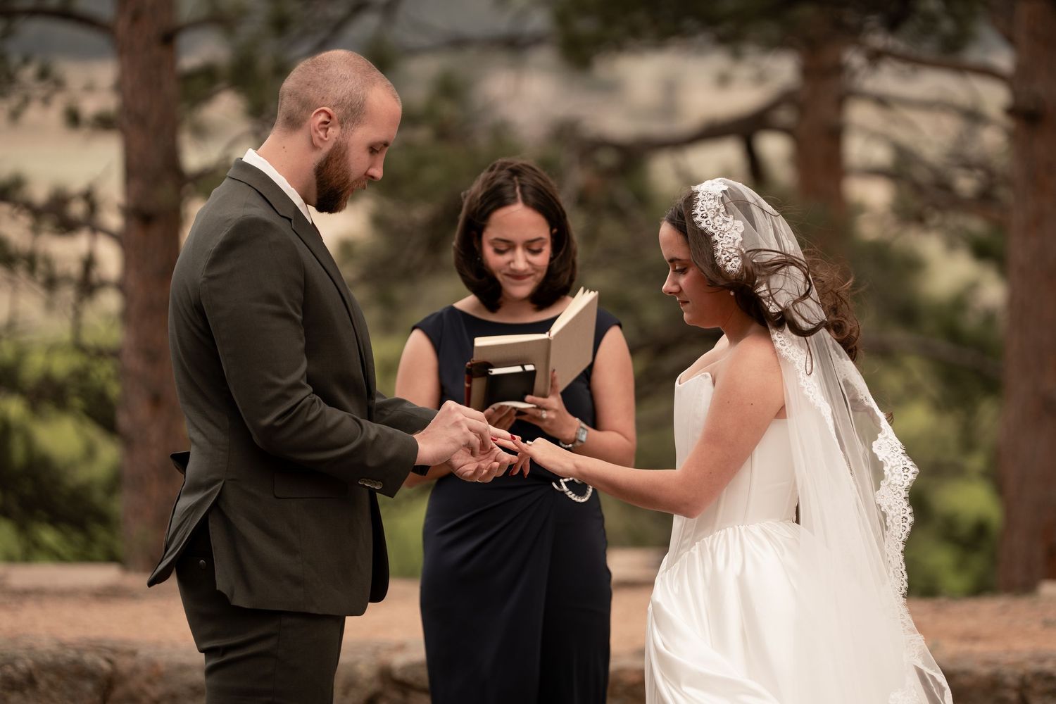 Bride and groom exchange rings during outdoor wedding ceremony with officiant in black dress, surrounded by pine trees.
