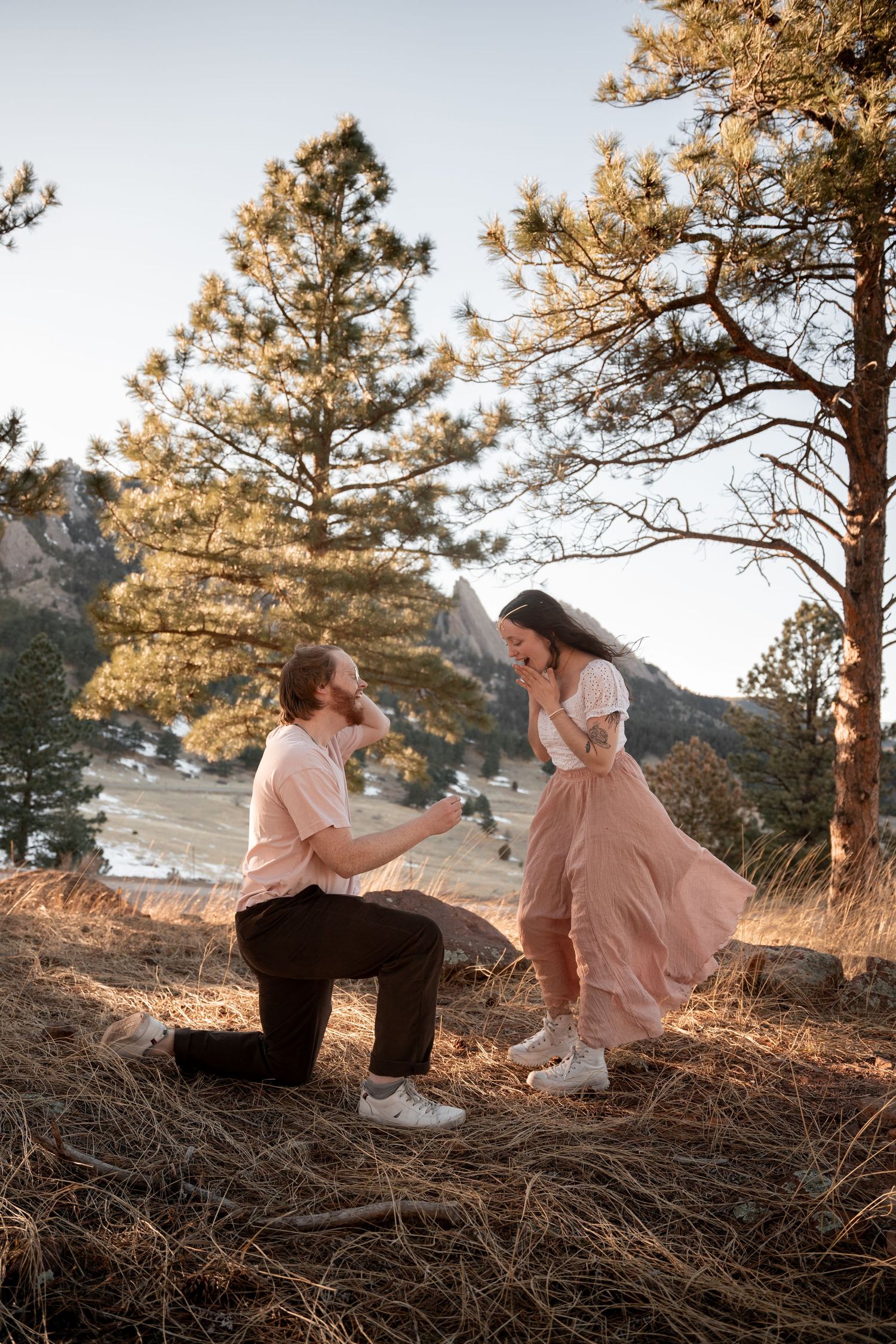 Man proposes on one knee to surprised woman in pink skirt among pine trees with mountain backdrop at golden hour.