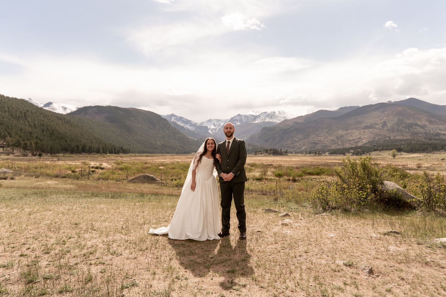 Bride and groom pose in mountain meadow with Rocky Mountain peaks and pine forests backdrop on their wedding day.