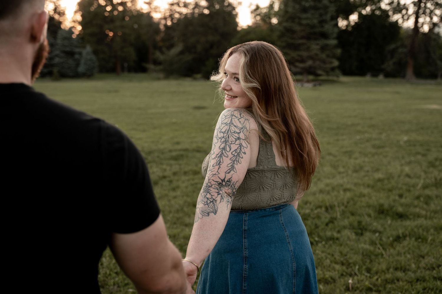 A smiling woman with a floral arm tattoo holds hands with a man in a black shirt in a sunlit green park.
