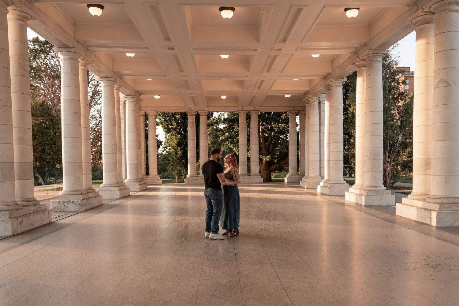 Couple embracing under elegant marble colonnade with tall white columns at golden hour.