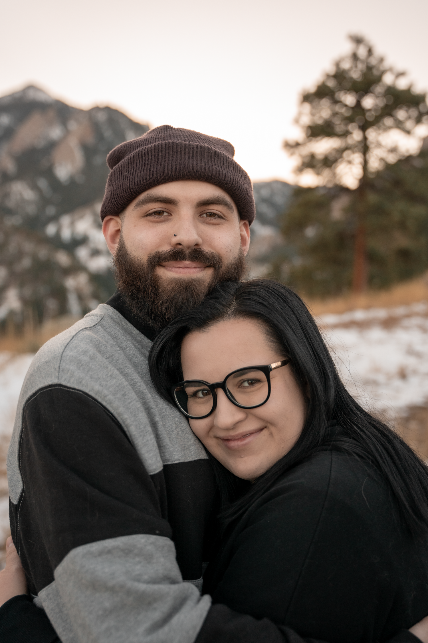 A cozy winter photoshoot featuring a couple embracing in warm clothing against a snowy mountain backdrop.