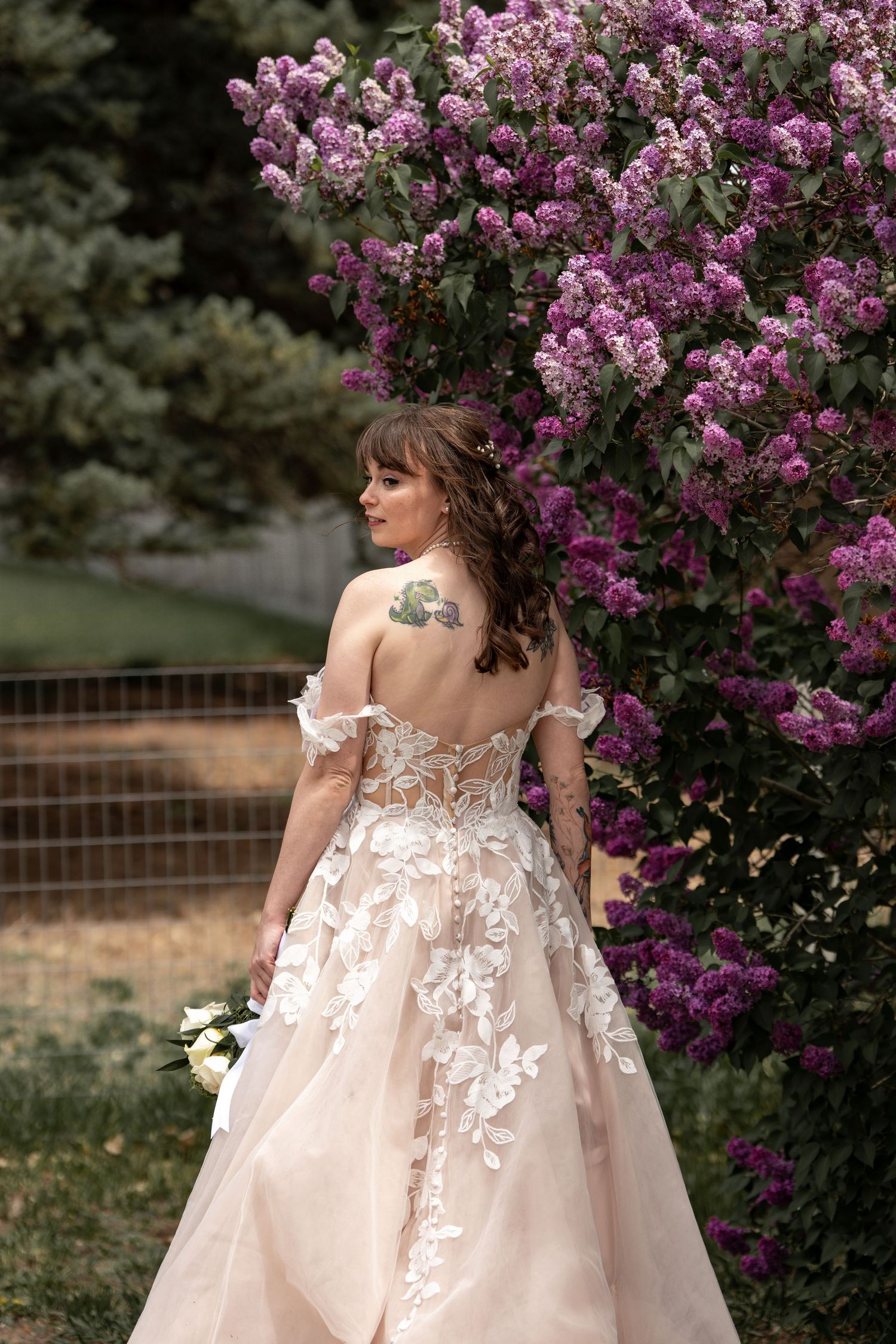 Bride in delicate lace gown poses near blooming purple lilac bushes in garden setting.