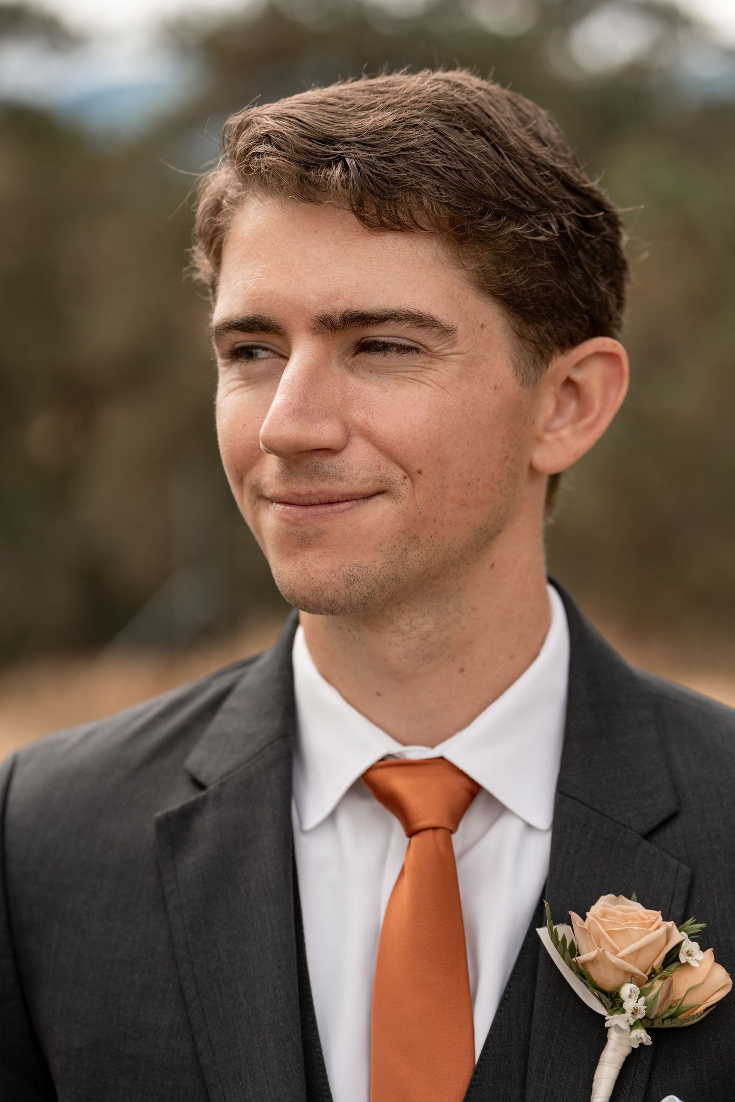 Groom wearing a dark suit with orange tie and peach boutonniere smiling at the camera.