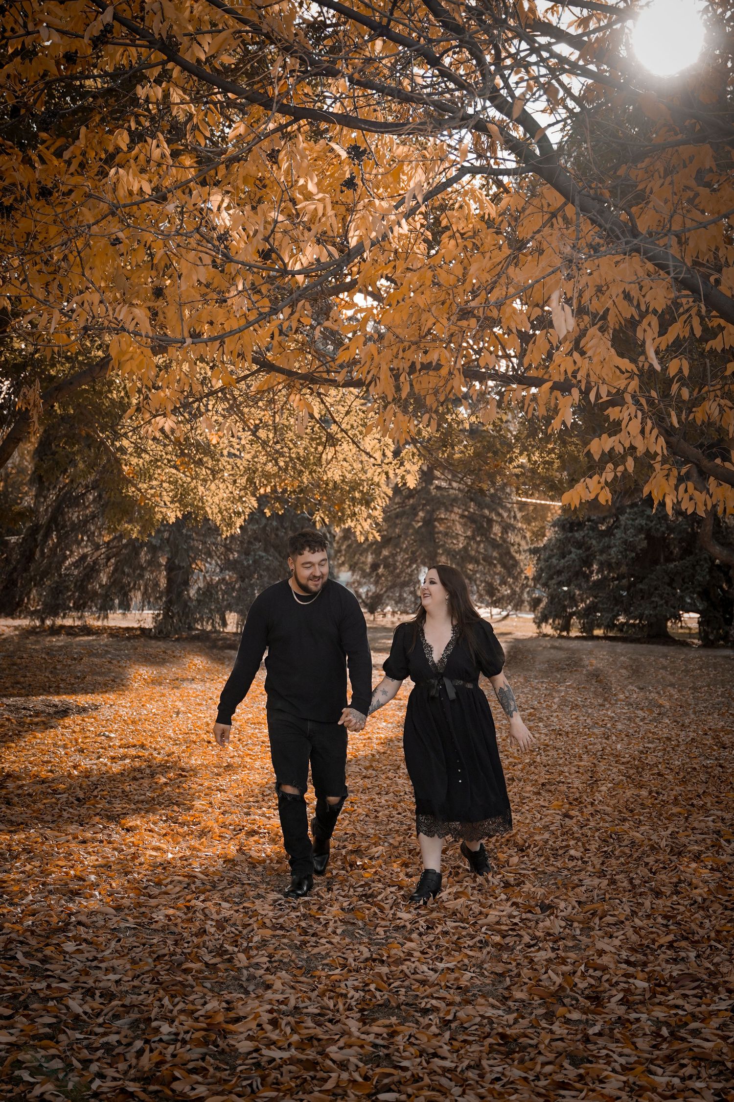 A couple walks together through a park filled with golden autumn leaves on a sunny afternoon.