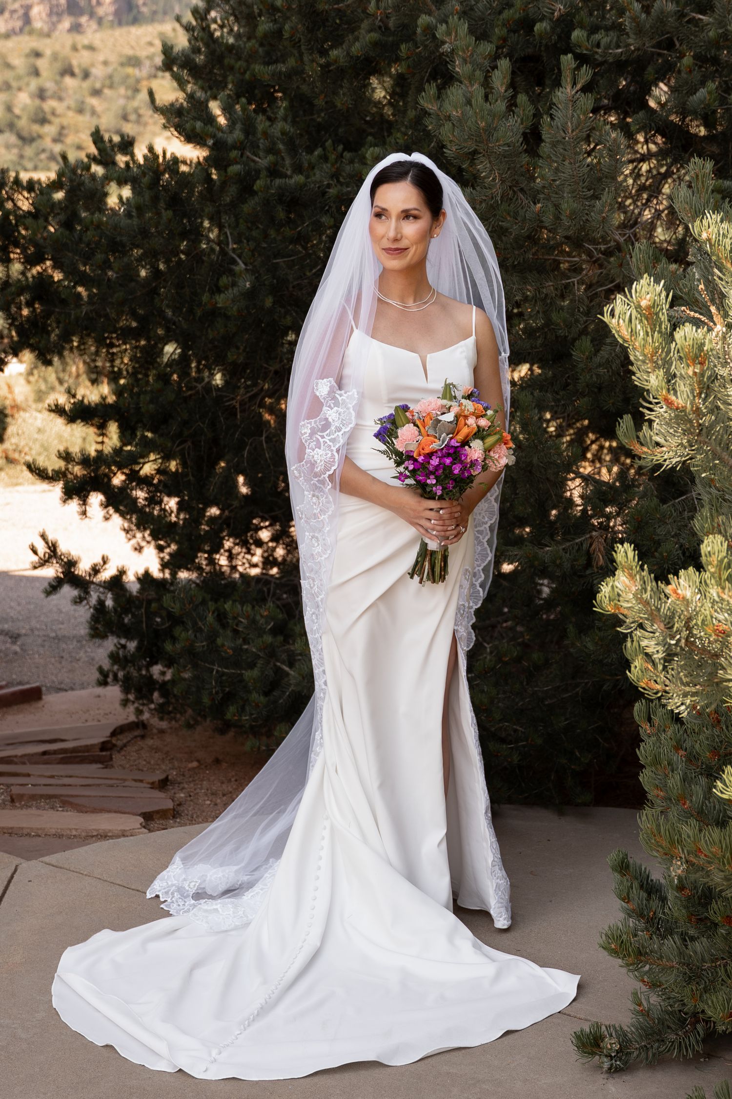 Elegant bride in fitted white gown holding colorful bouquet against natural backdrop.