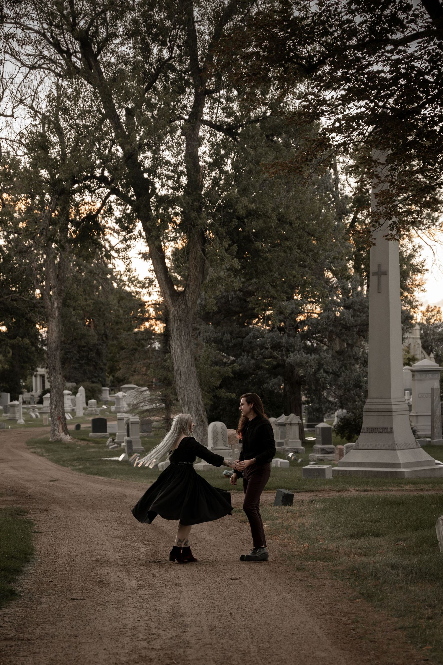 Couple in black clothing dancing playfully in a cemetery at sunset, surrounded by gravestones and trees.