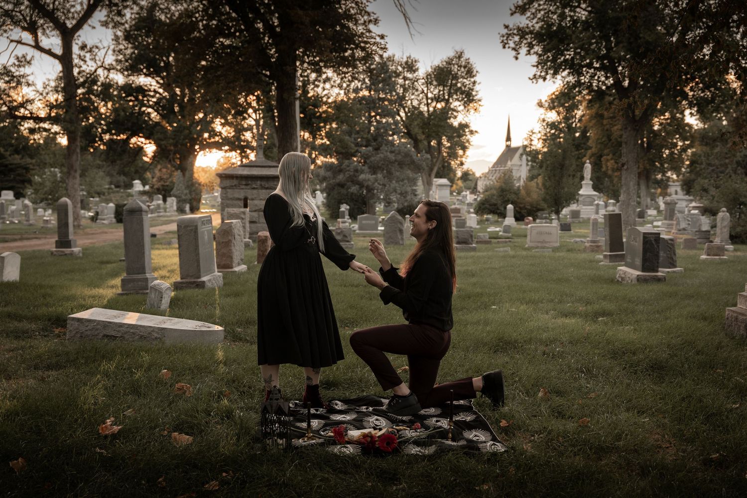 A man proposes to a woman on a blanket in a cemetery at sunset, surrounded by gravestones and rose petals.