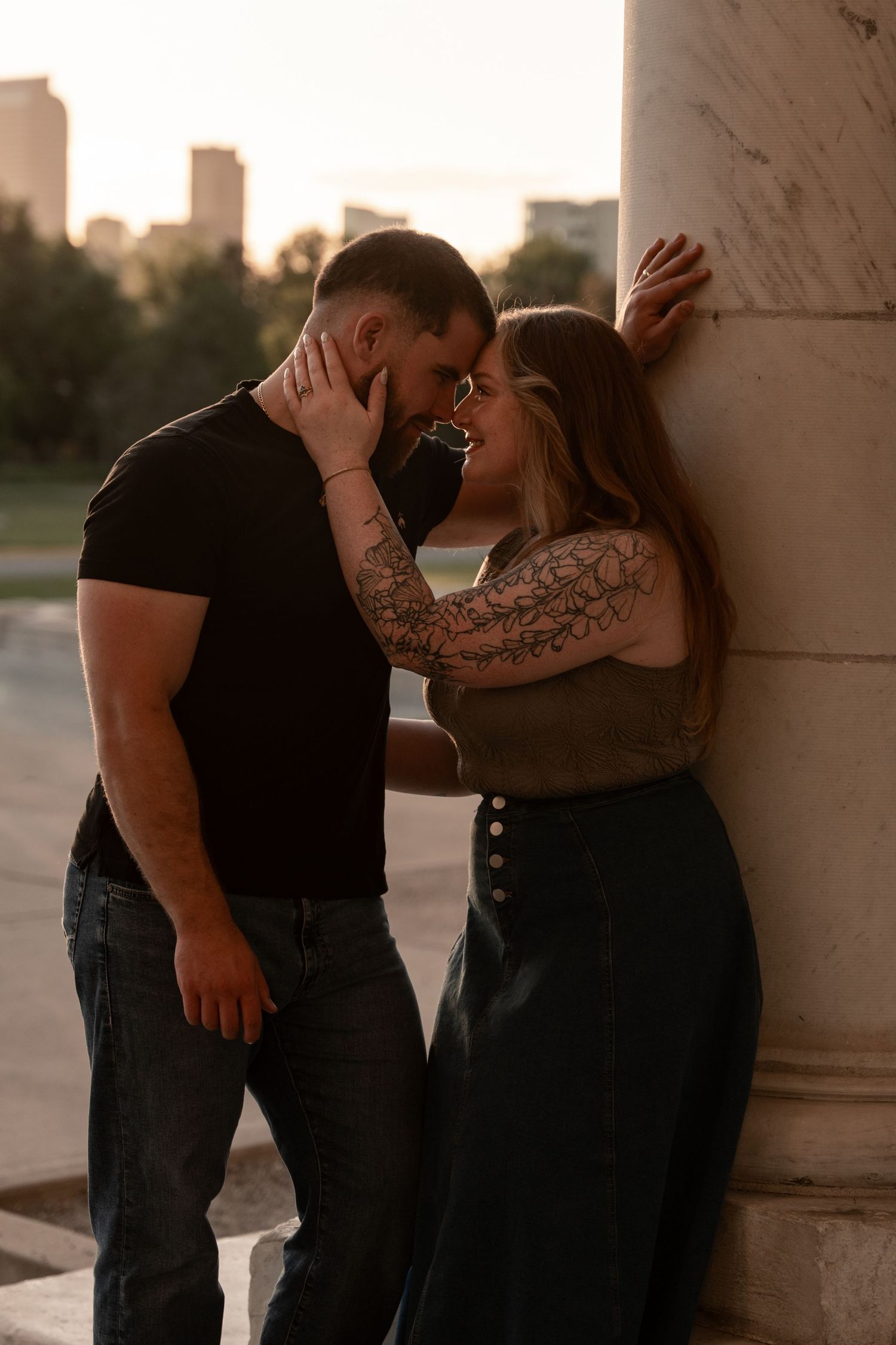Couple with tattoos sharing a romantic moment leaning against a marble column at golden hour with city skyline backdrop.