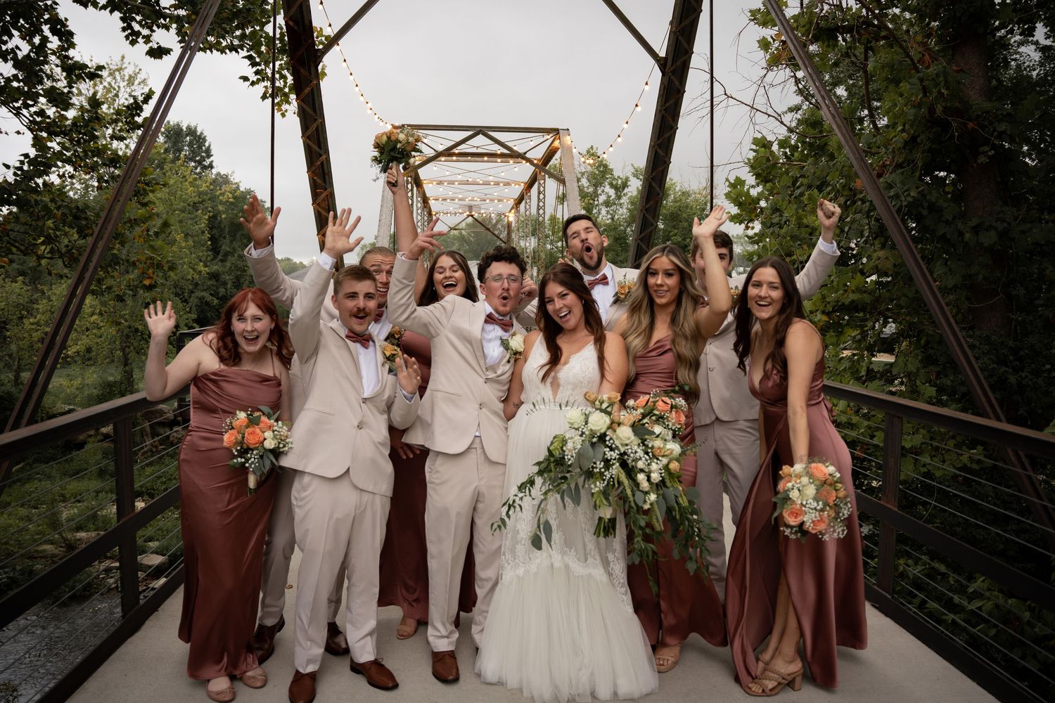A wedding party celebrates joyfully on a rustic metal bridge surrounded by lush greenery.