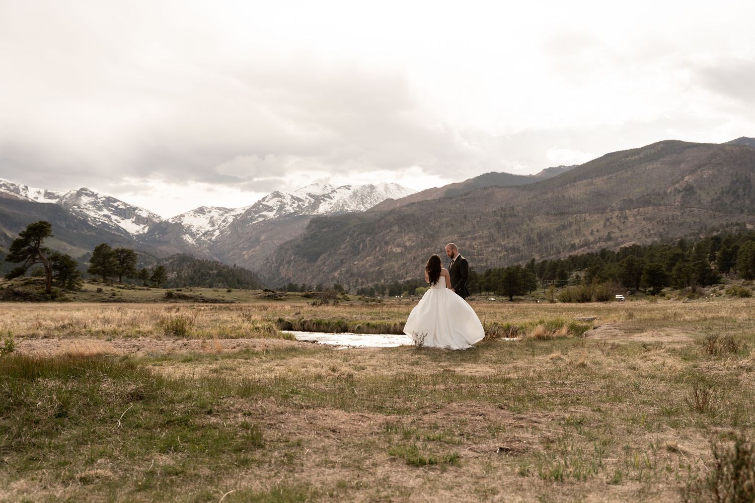 Bride and groom stand together gazing at snow-capped Rocky Mountains in a vast meadow on their wedding day.