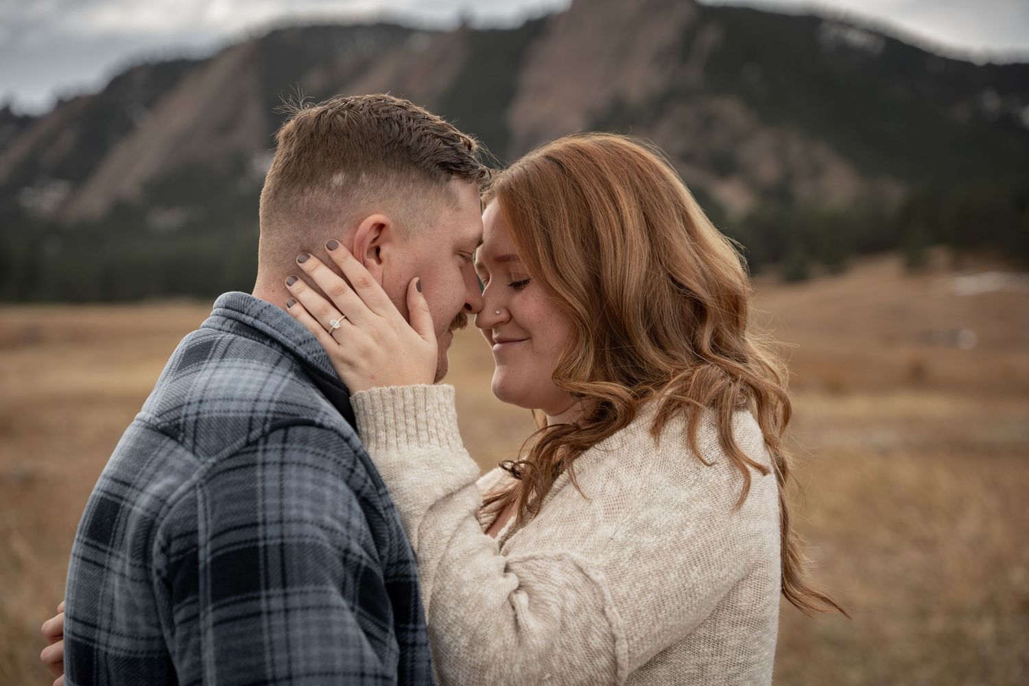 Couple sharing a romantic moment in a mountain field, touching foreheads and smiling, wearing cozy fall outfits.