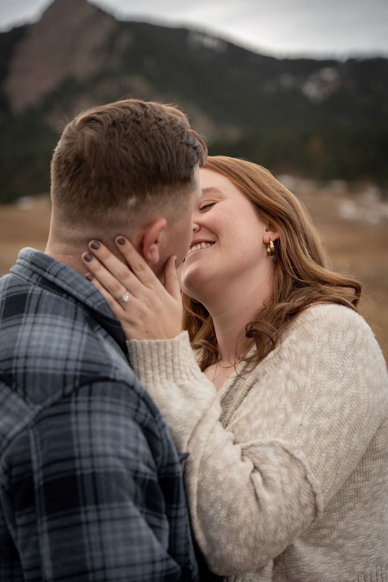 A joyful couple shares a romantic moment outdoors near mountains, woman in cream sweater smiling as they lean in for a kiss.