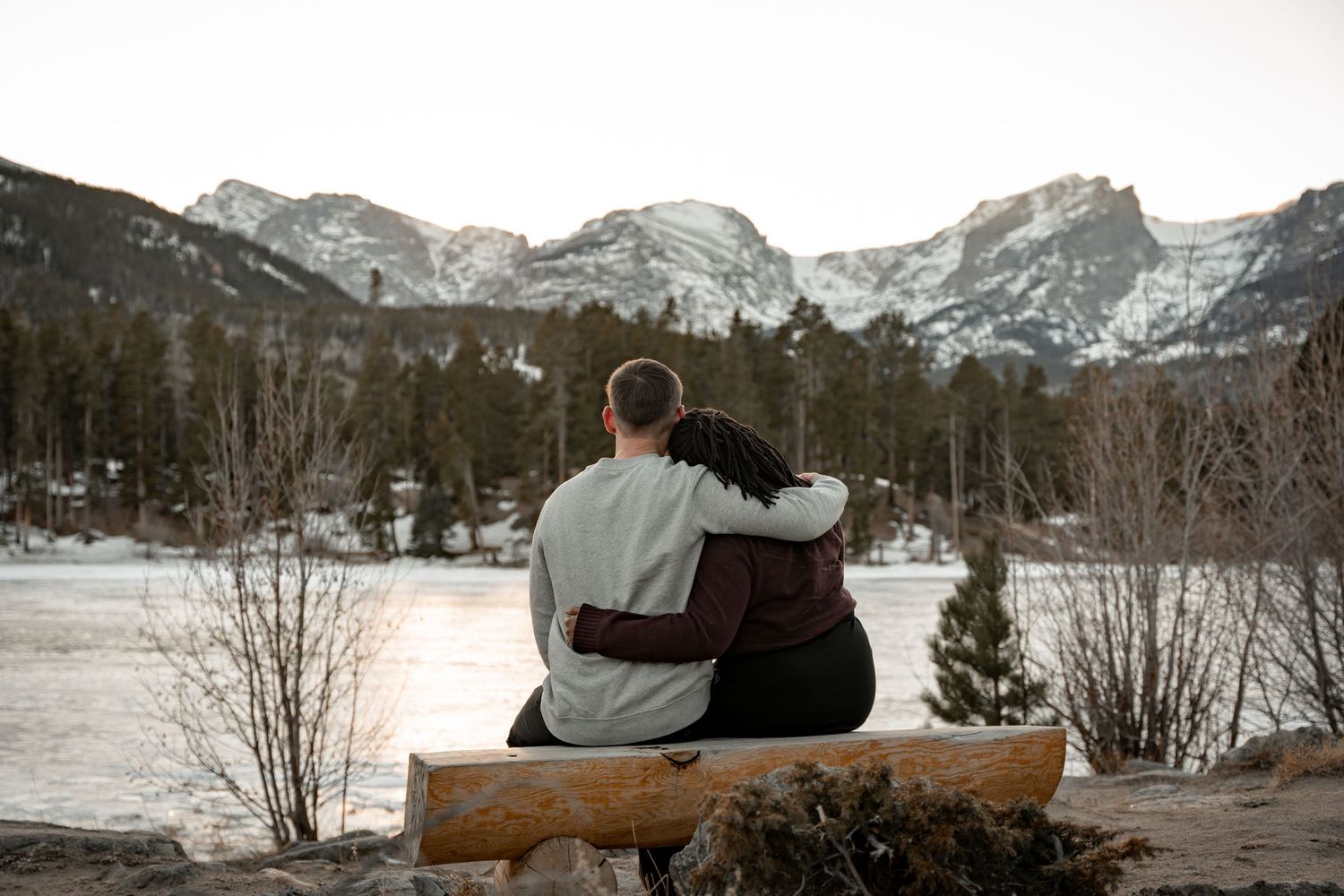 Couple embraces on a log bench overlooking a frozen mountain lake surrounded by snow-dusted pine trees.