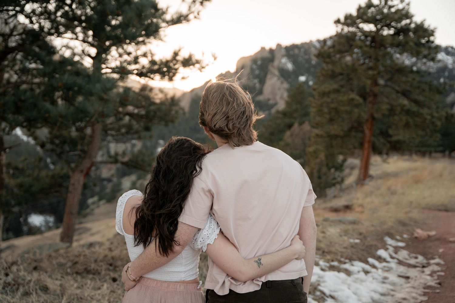 Couple embracing while watching sunset over mountain landscape surrounded by pine trees.