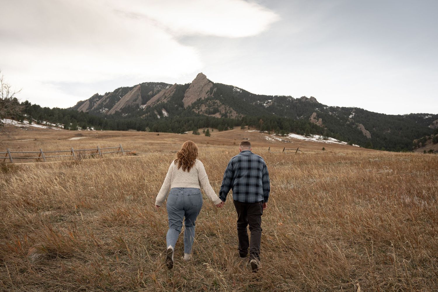 Couple holding hands walking through golden field with rocky Flatirons mountains in background, Boulder Colorado.