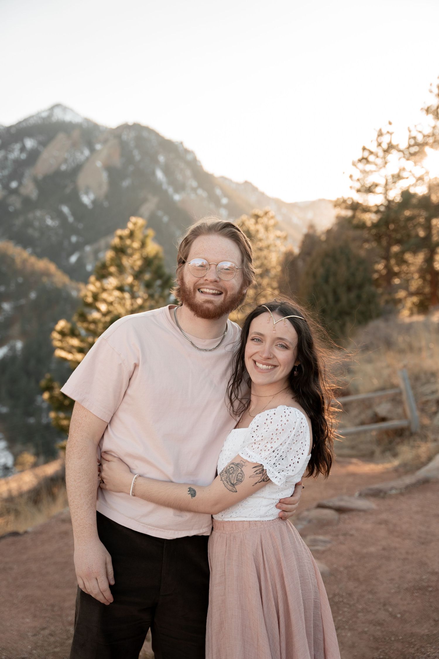 A smiling couple embraces outdoors with a stunning mountain backdrop and golden sunset light filtering through pine trees.