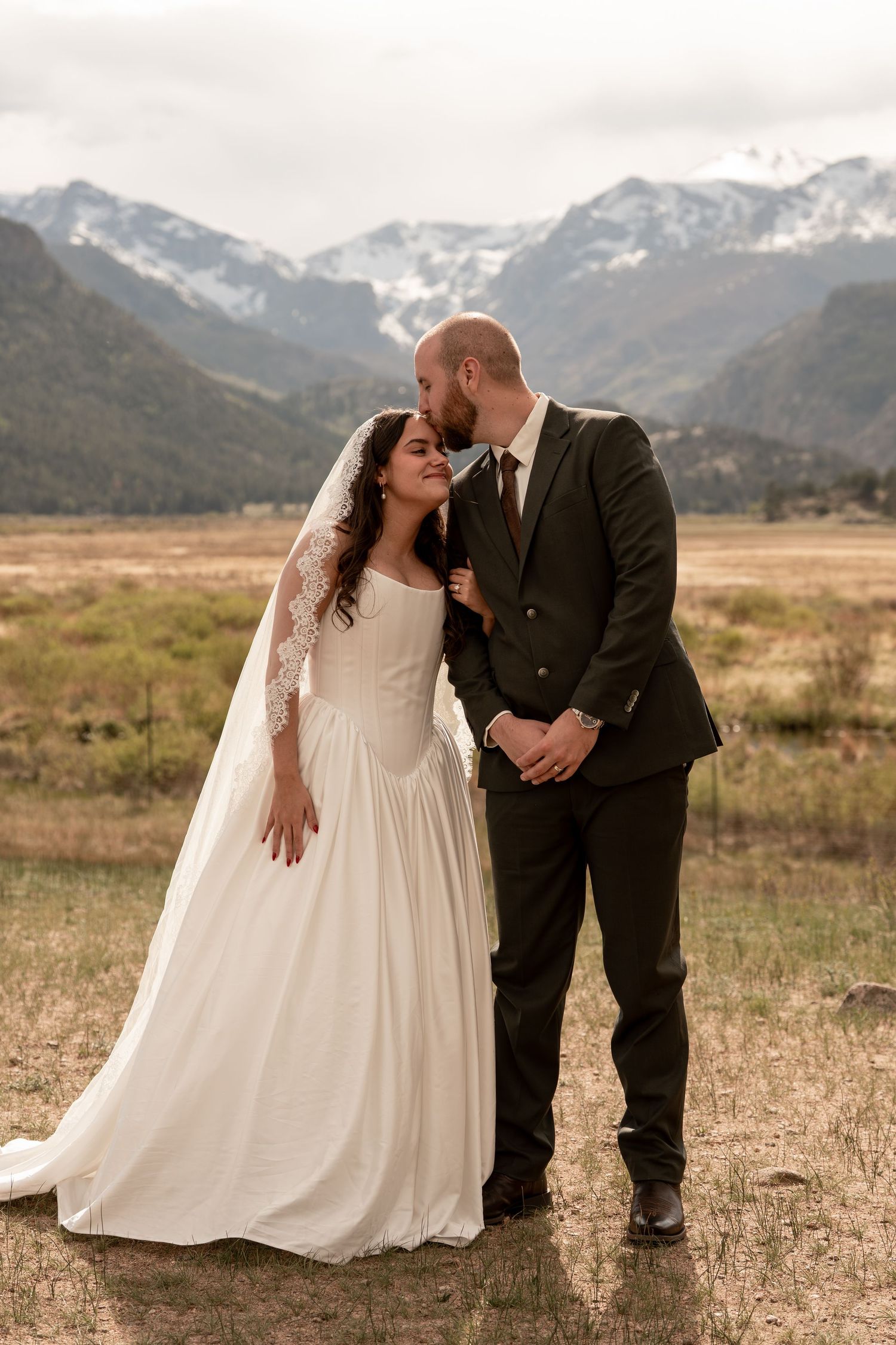 Bride and groom embracing in a mountain meadow, with snow-capped Rocky Mountains in the background.