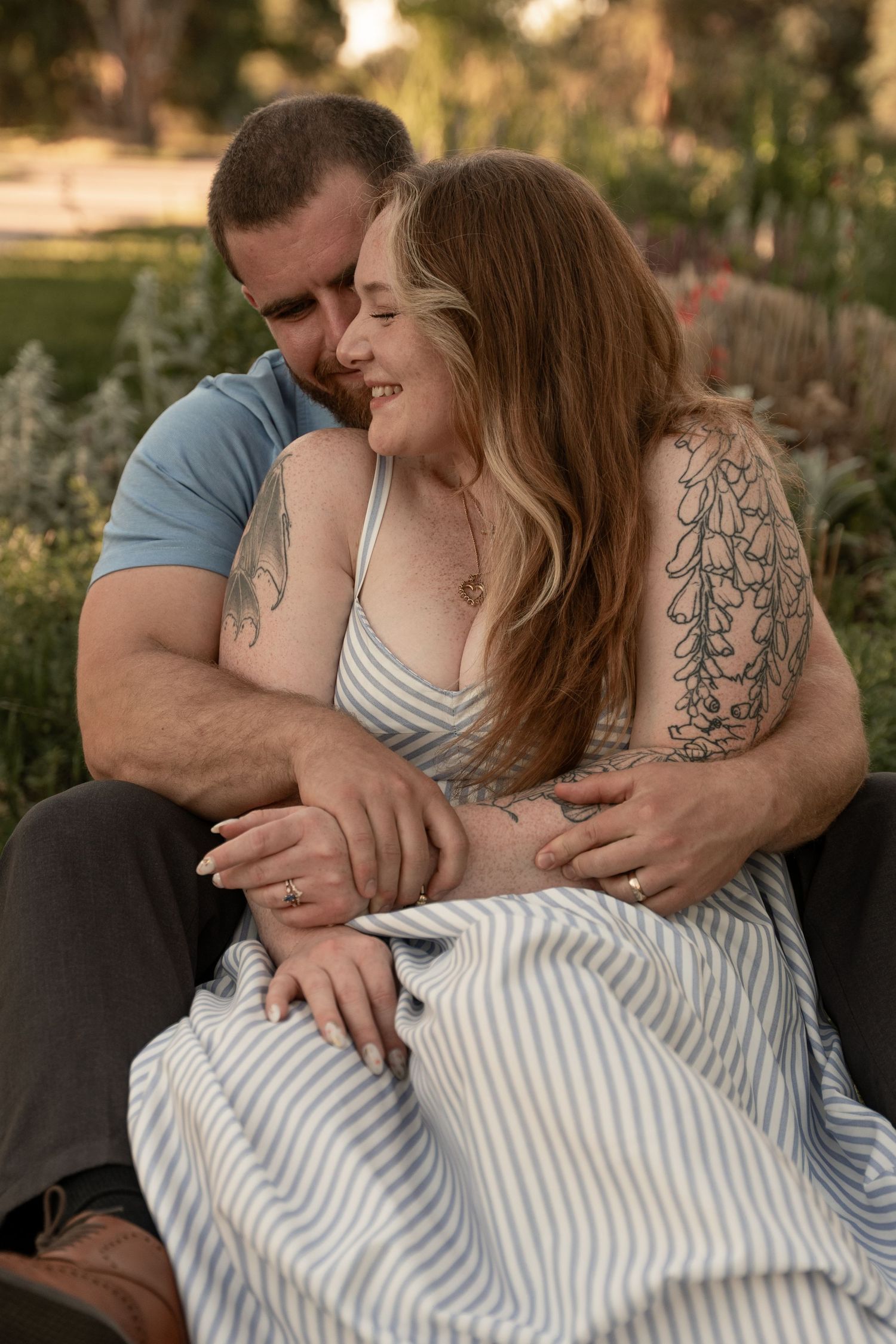 A couple sits together outdoors, the man embracing the tattooed woman in a striped dress as she smiles warmly in a garden setting.