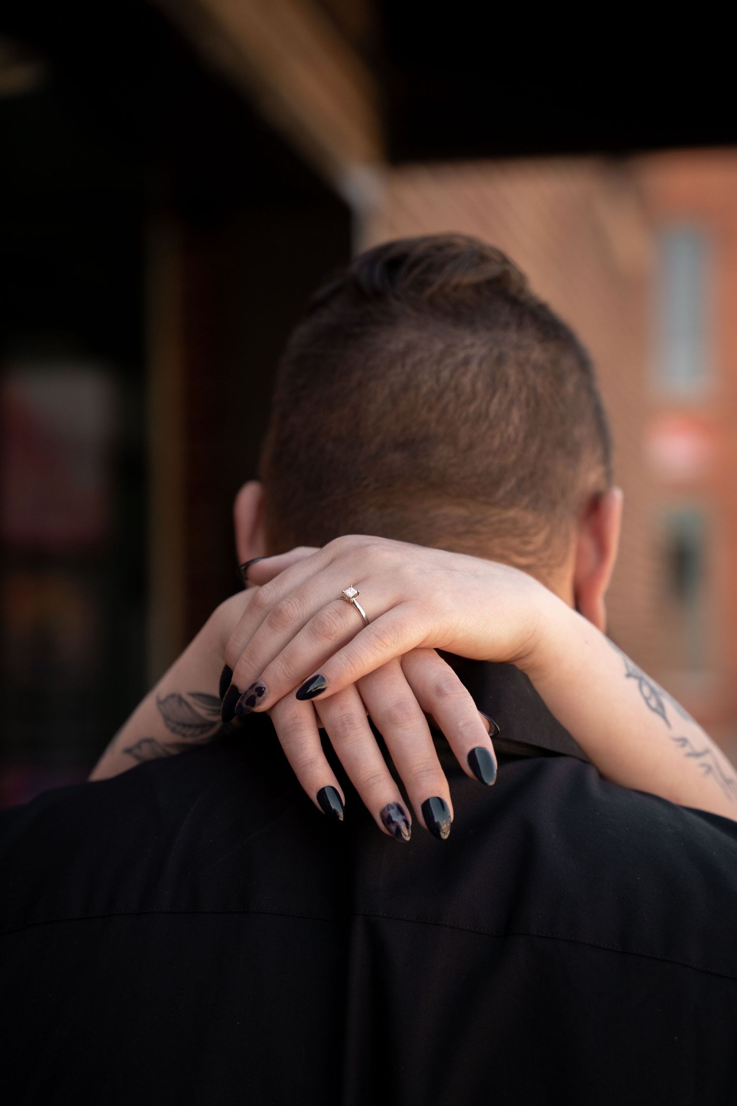 Close-up of tattooed woman's hand with engagement ring and black nails resting on man's shoulder.