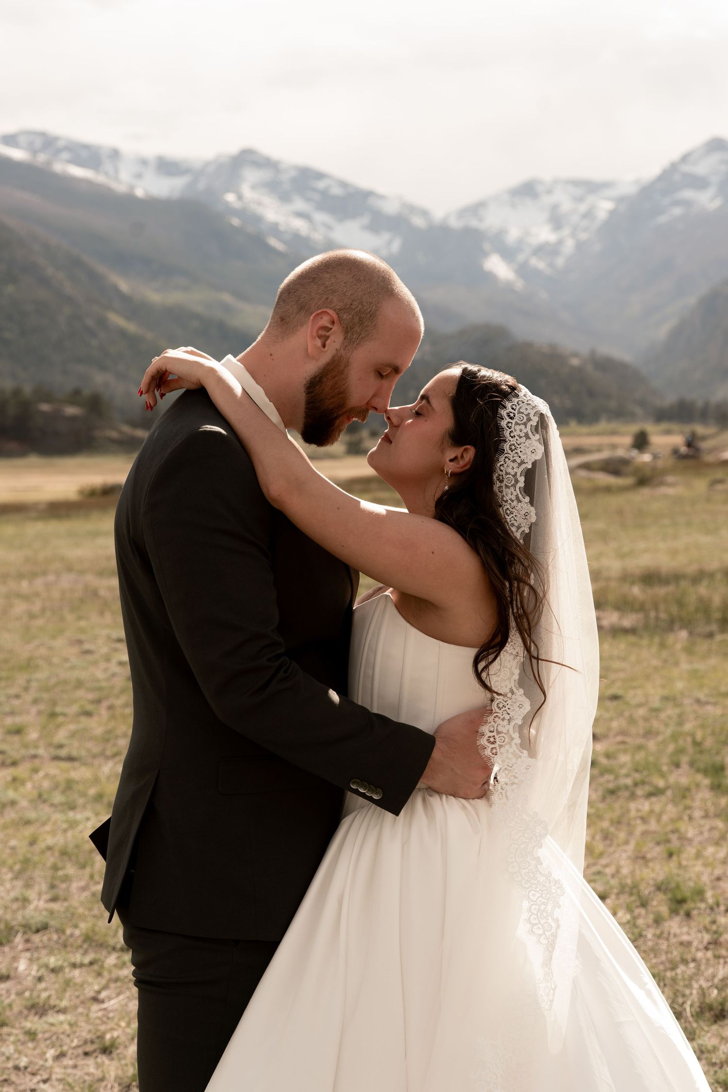 Bride and groom embracing in a mountain meadow, wearing wedding attire with snow-capped peaks in the background.