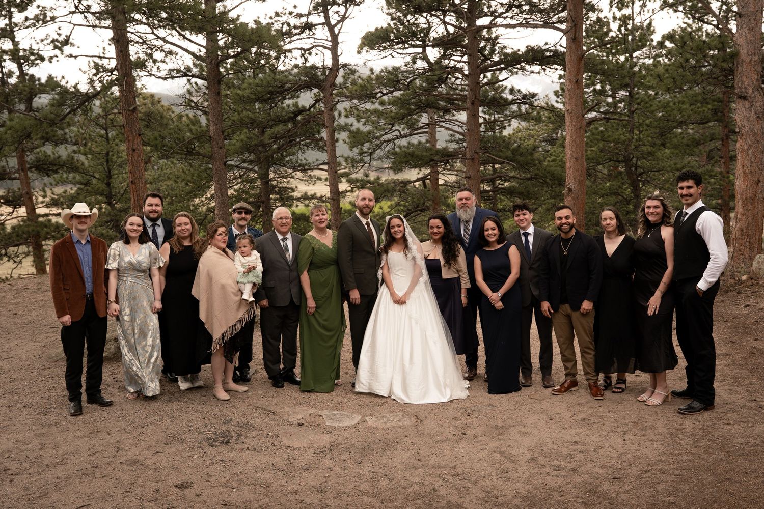 Wedding party poses outdoors among pine trees, bride in white gown surrounded by family and friends in formal attire.
