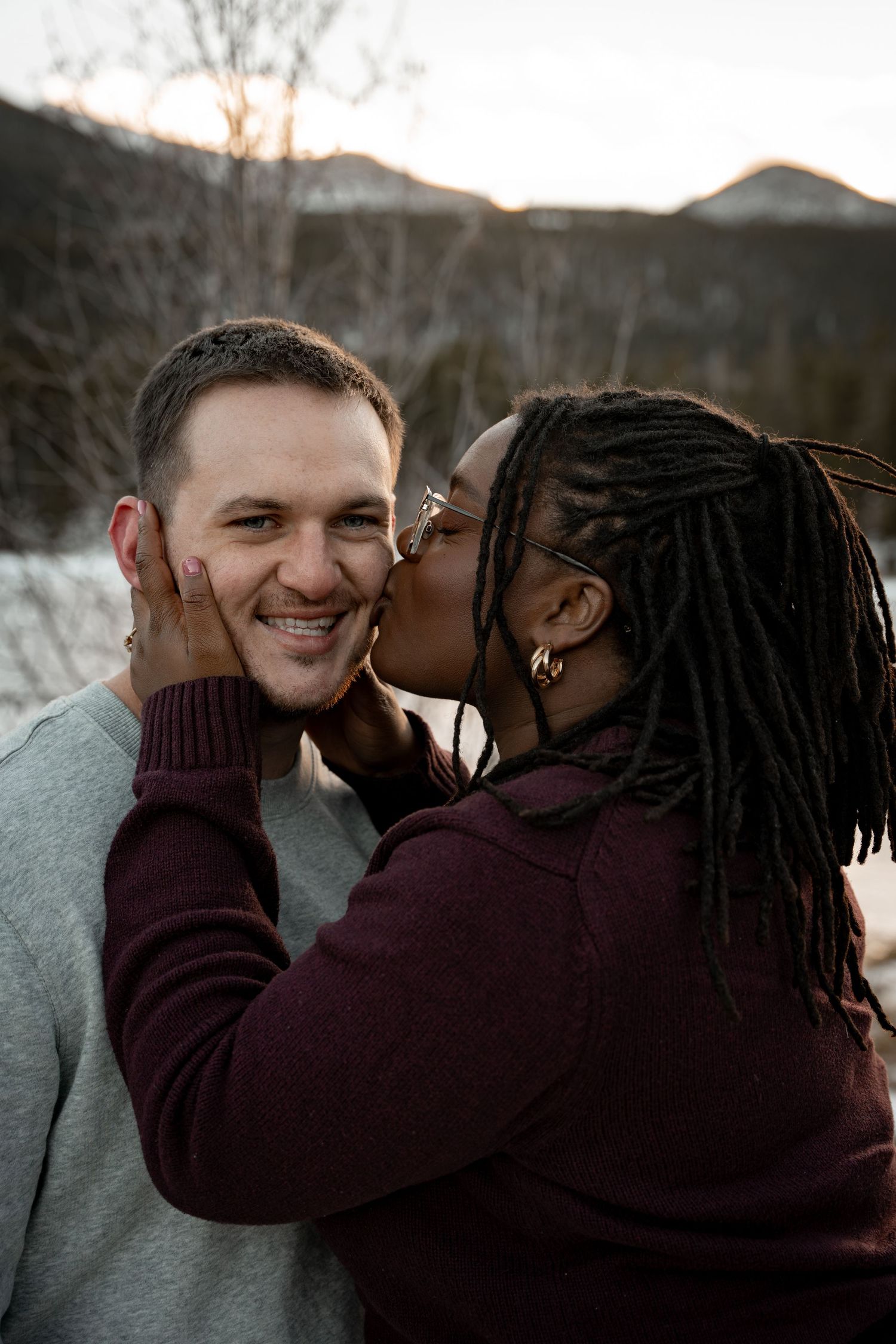 Woman with dreadlocks kissing smiling man on cheek with snowy mountains in background.