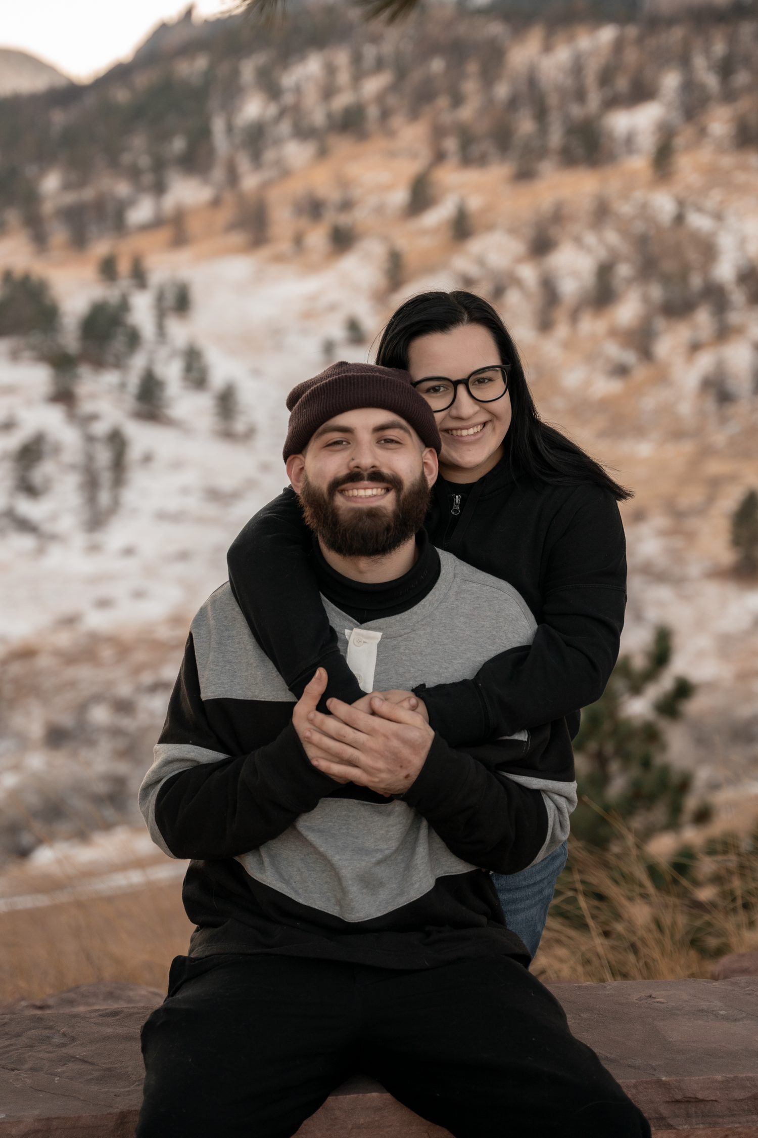 A smiling couple poses outdoors in winter, woman hugging man from behind, with snowy mountain landscape behind them.