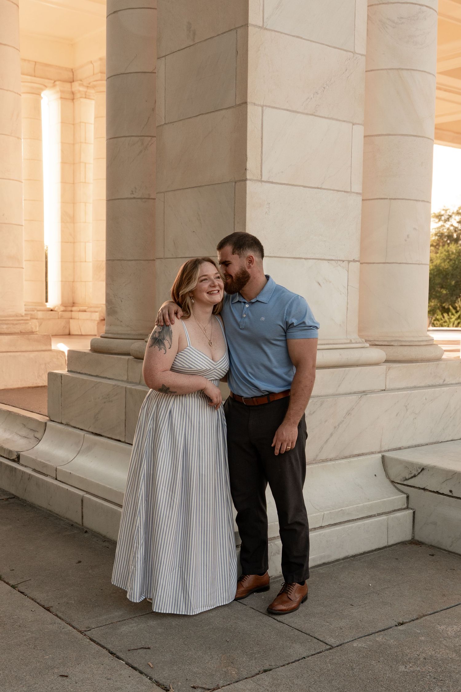 Couple embracing and smiling beside marble columns at golden hour, woman in striped maxi dress, man in blue polo shirt.