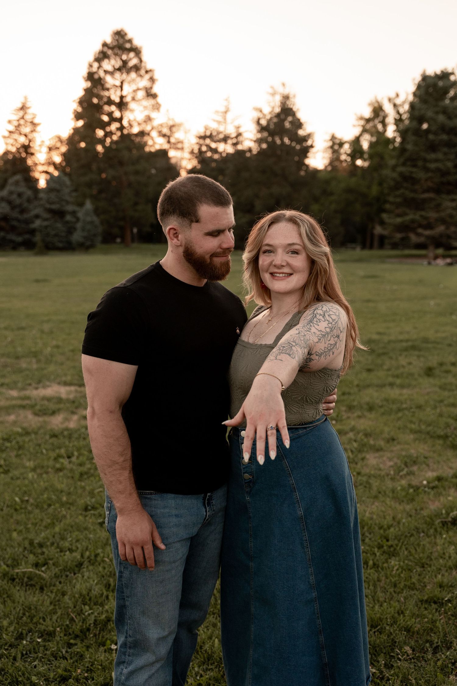Couple posing at golden hour in a park, woman showing engagement ring, man in black tee and jeans, trees in background.