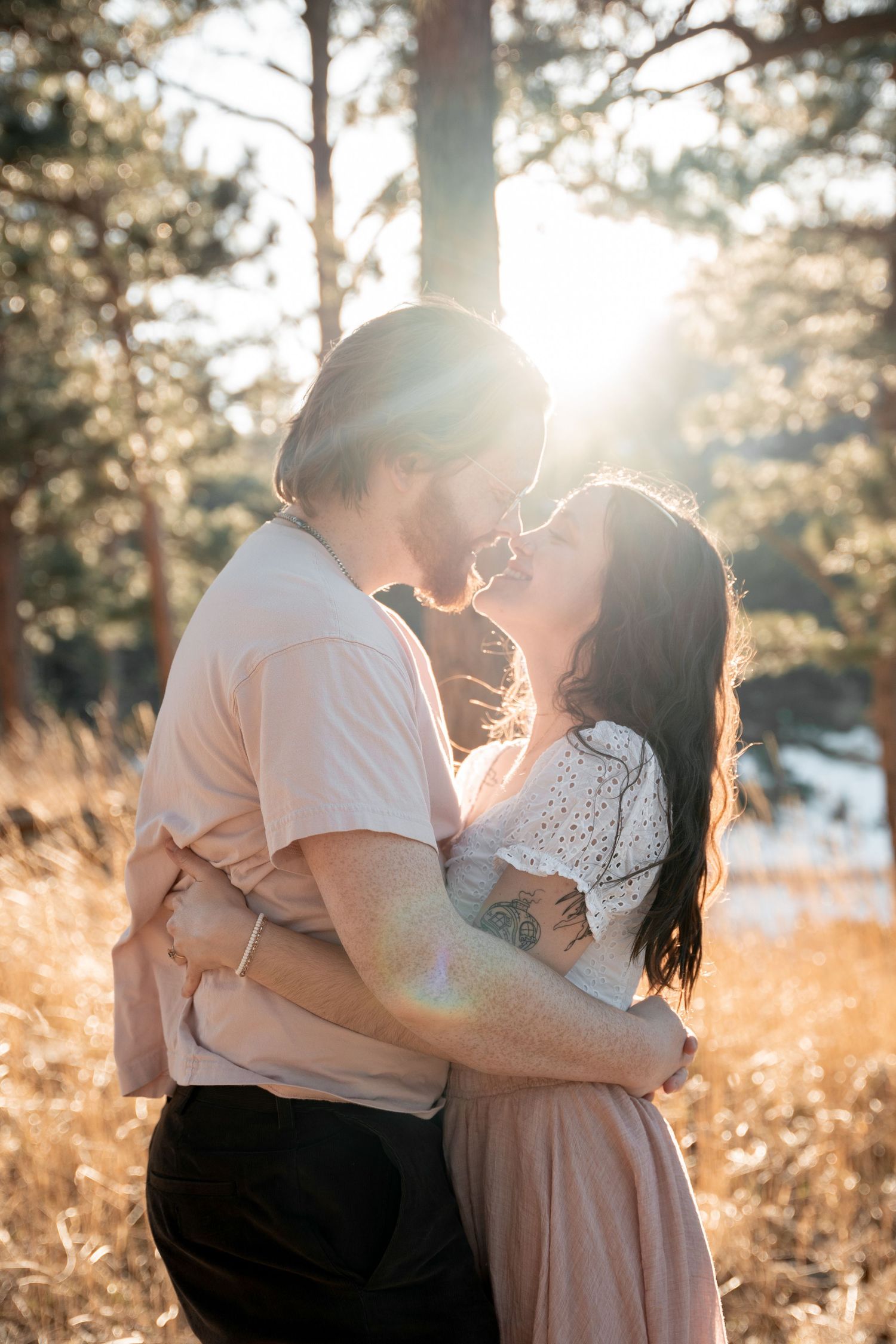 Couple sharing a romantic kiss in a sunlit forest clearing, surrounded by golden grass and pine trees.