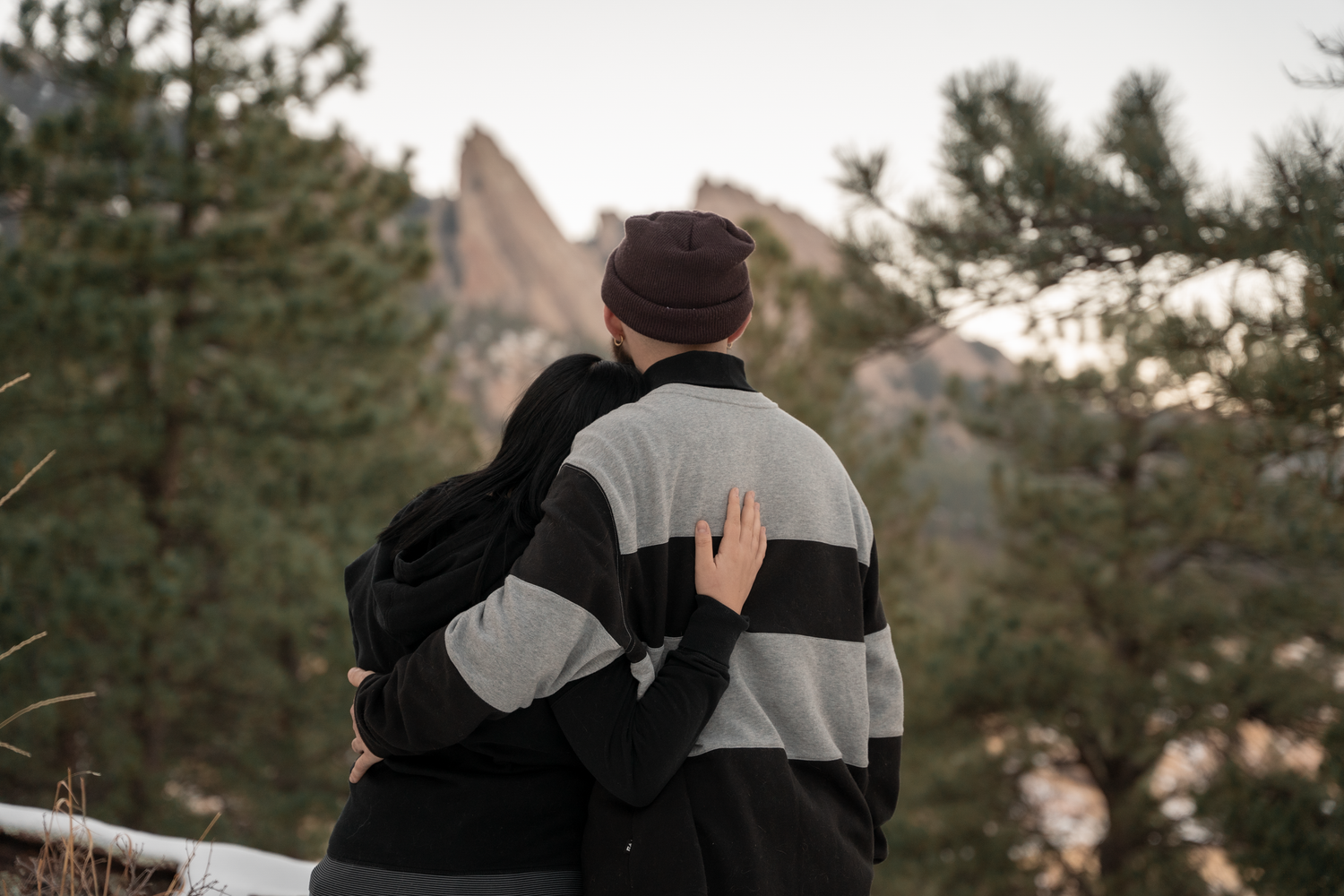 Couple embracing from behind admiring rocky mountain landscape surrounded by pine trees in winter.