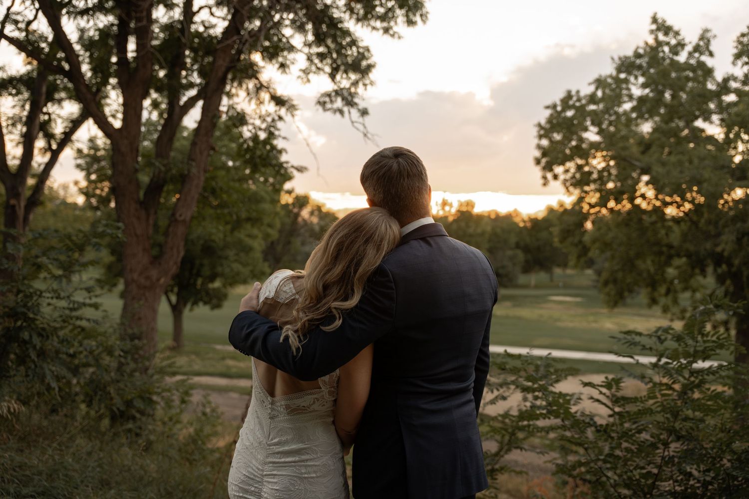 Newlyweds embrace while watching sunset in a scenic outdoor setting.