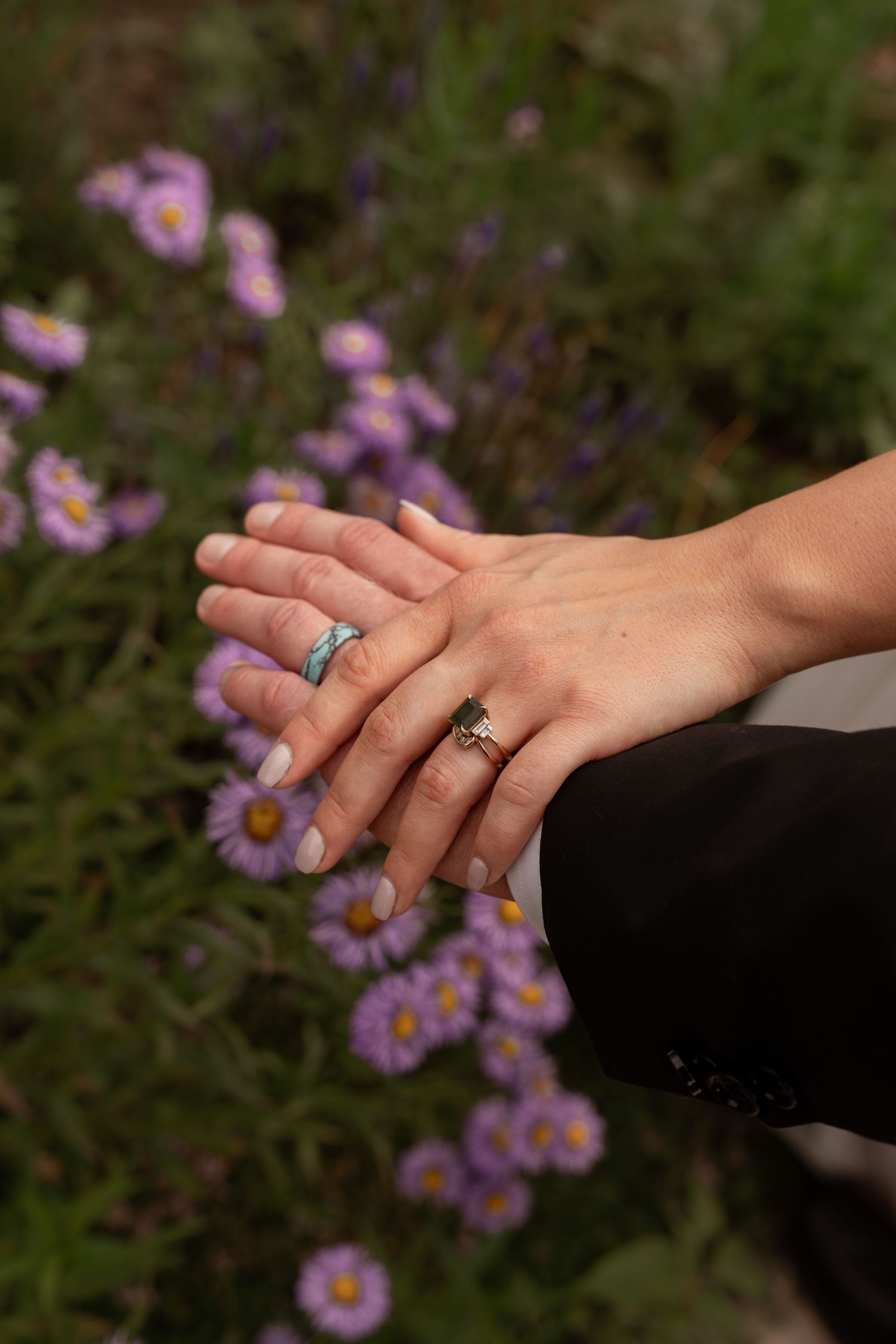 Two hands wearing engagement rings rest gently against a backdrop of purple wildflowers in a romantic outdoor setting.