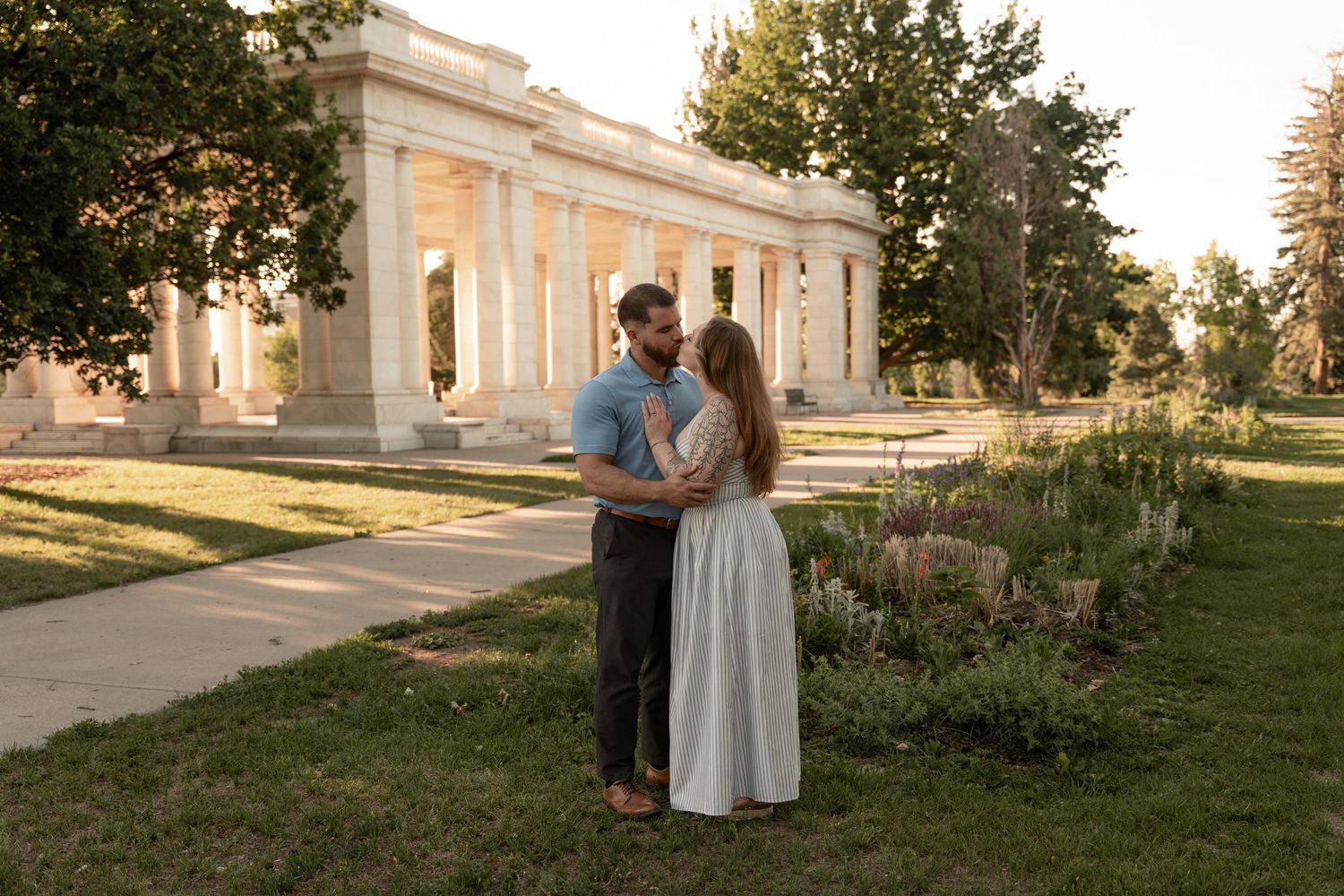Couple embracing in a lush green park near a white neoclassical colonnade during golden hour.