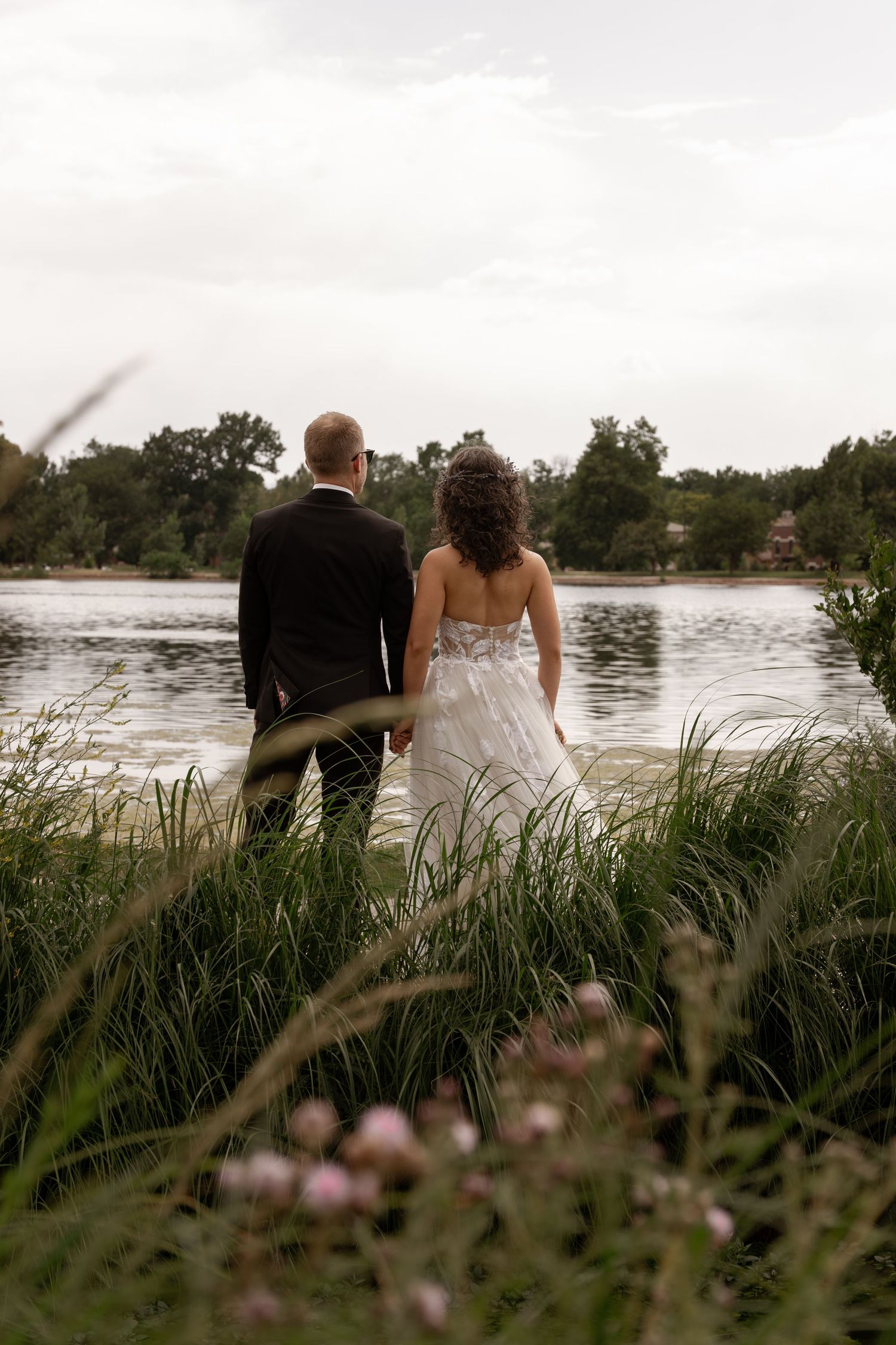 A couple sits by a tranquil lake surrounded by tall grass and wildflowers at sunset.