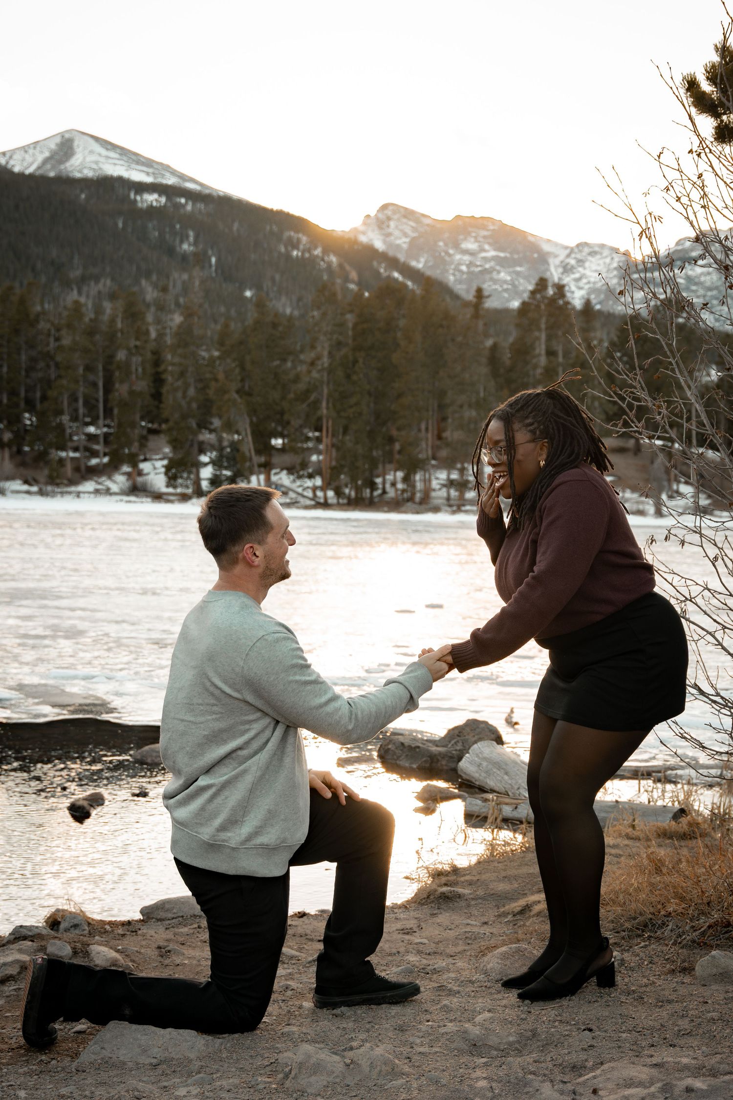 Man proposing on one knee to surprised woman by frozen mountain lake at sunset.
